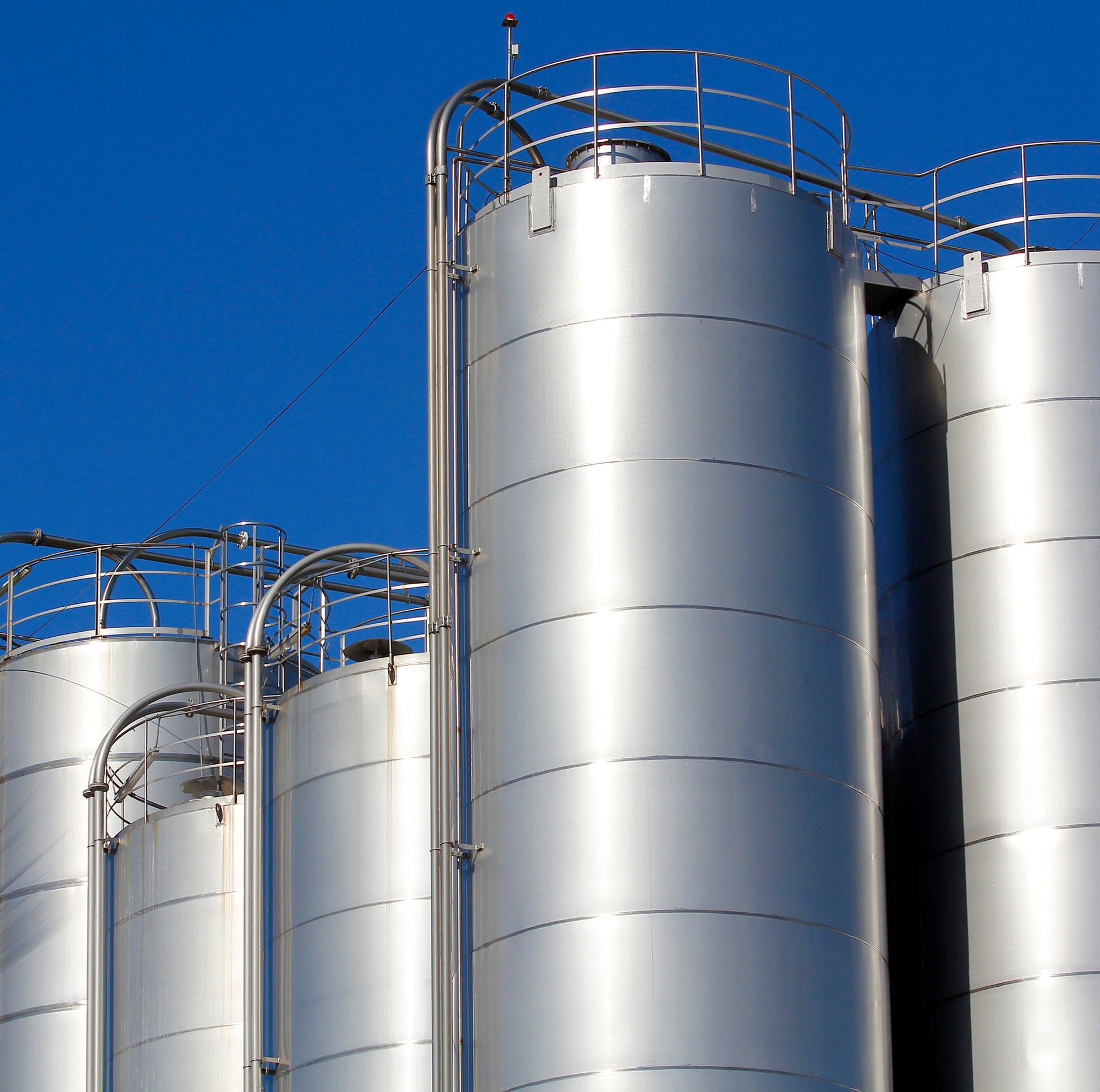 <Five large outdoor stainless steel silo tanks for receiving and storing liquid materials from tanker lorries on a bright sunny day with blue sky. Credit> AdobeStock_102652410