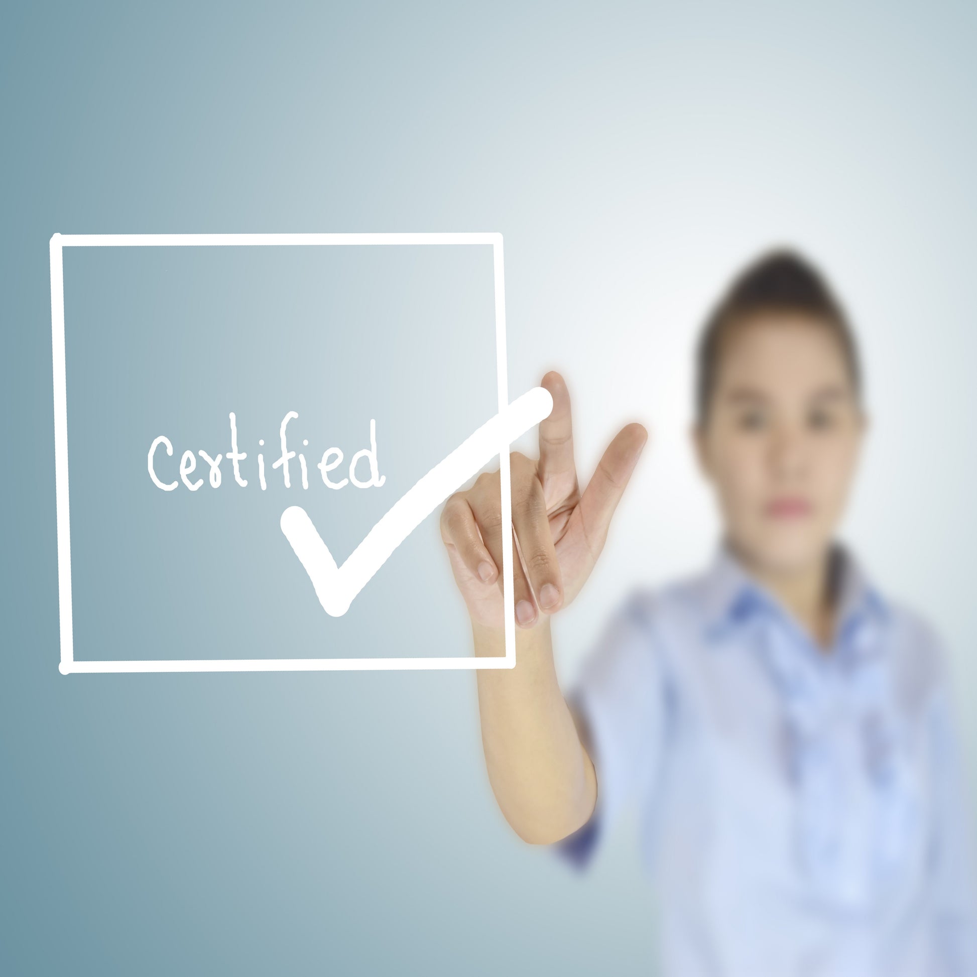 <Blurred woman with dark hair wearing a blue shirt pointing at a clear screen to tick a white lined box containing the word certified for approval. Credit> AdobeStock_104109923