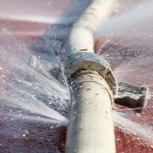 <Burst, white, 10cm diameter water pipe or hose crossing a sunlit, red concrete surface  leaking water in several places under pressure. Credit> AdobeStock_119492760