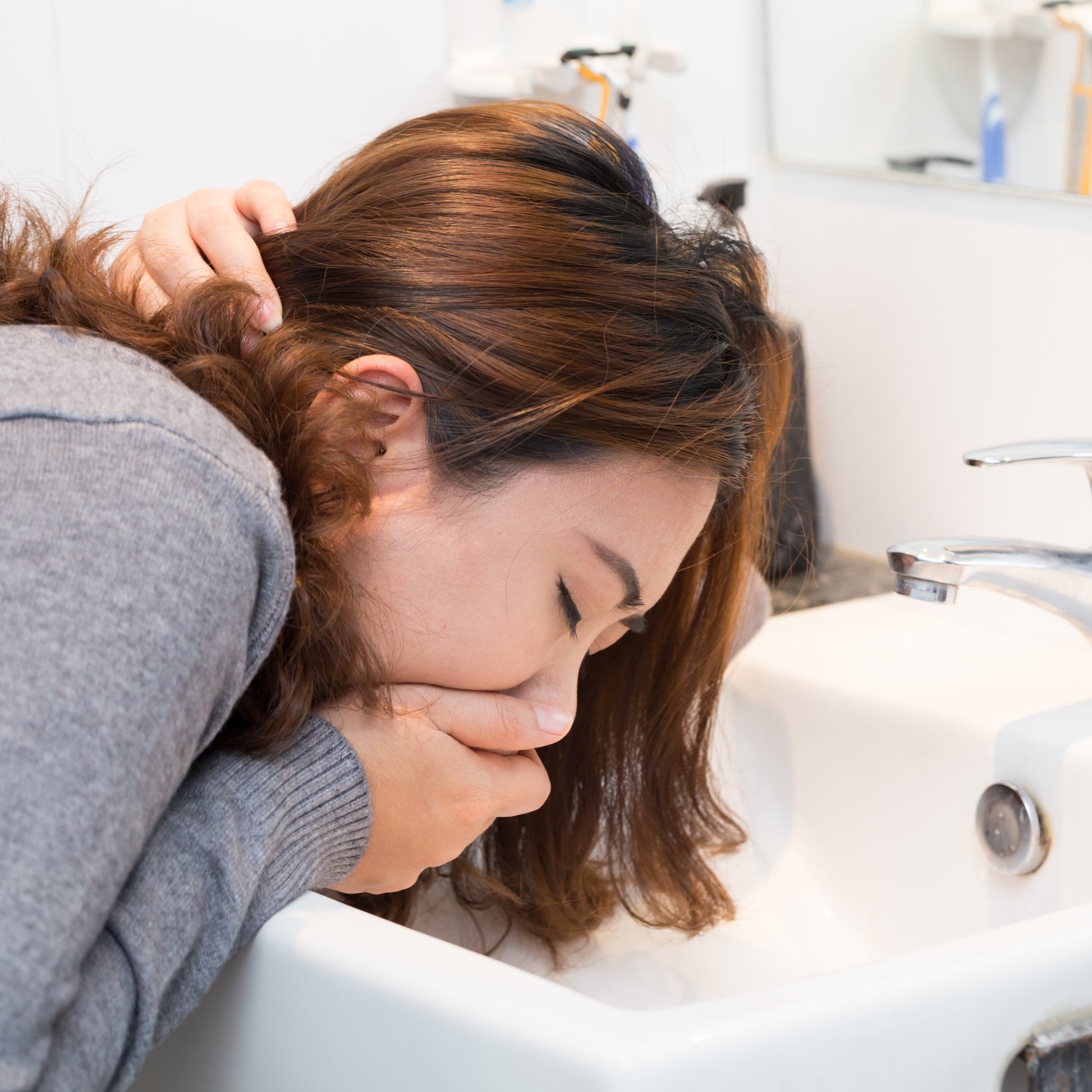 <Chinese female employee with auburn coloured hair, wearing a grey jumper, off work sick holding her mouth leaning into a white bathroom sink below a mirror about to vomit, Credit> AdobeStock_137810648