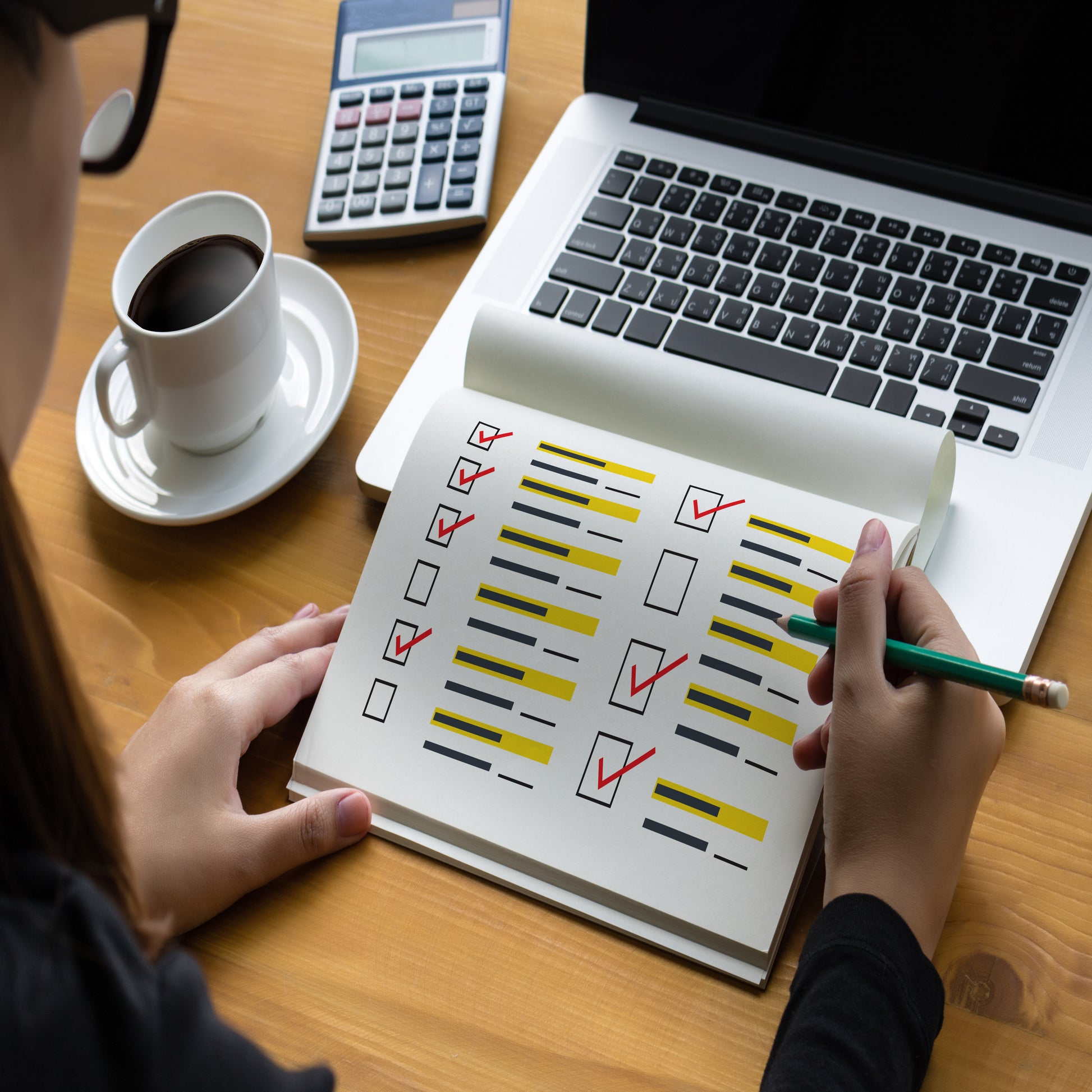 <Part face of a brunette woman wearing black glasses and a black shirt, completing a paper checklist sitting on a silver and black laptop keyboard next to a cup of coffee and a calculator. Credit> AdobeStock_168686189_1