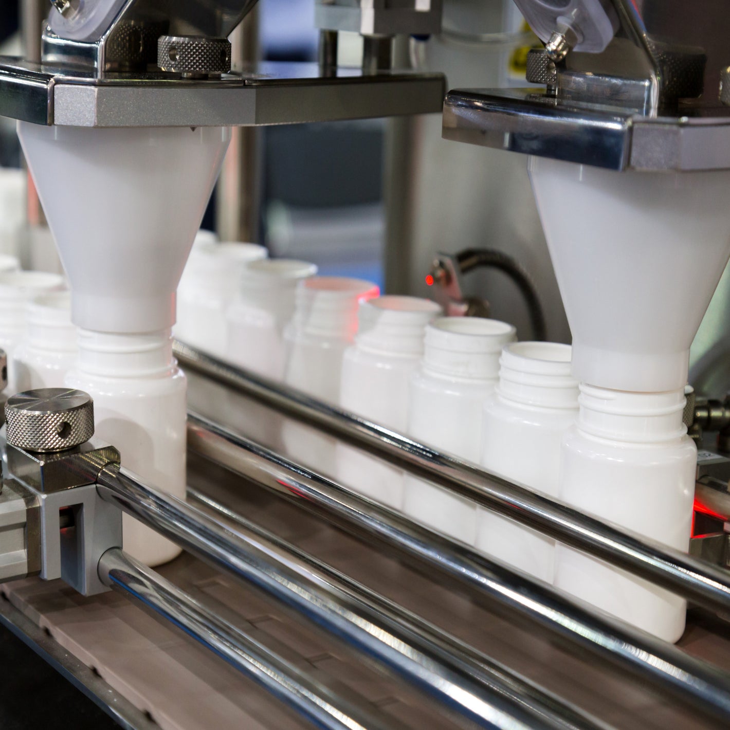 <Two rows of white bottles passing down a conveyor belt, held in place with two chrome circular guardrails on each side, being filled with dietary supplement capsules from a white funnel connected to a filling machine. Credit> AdobeStock_169195287