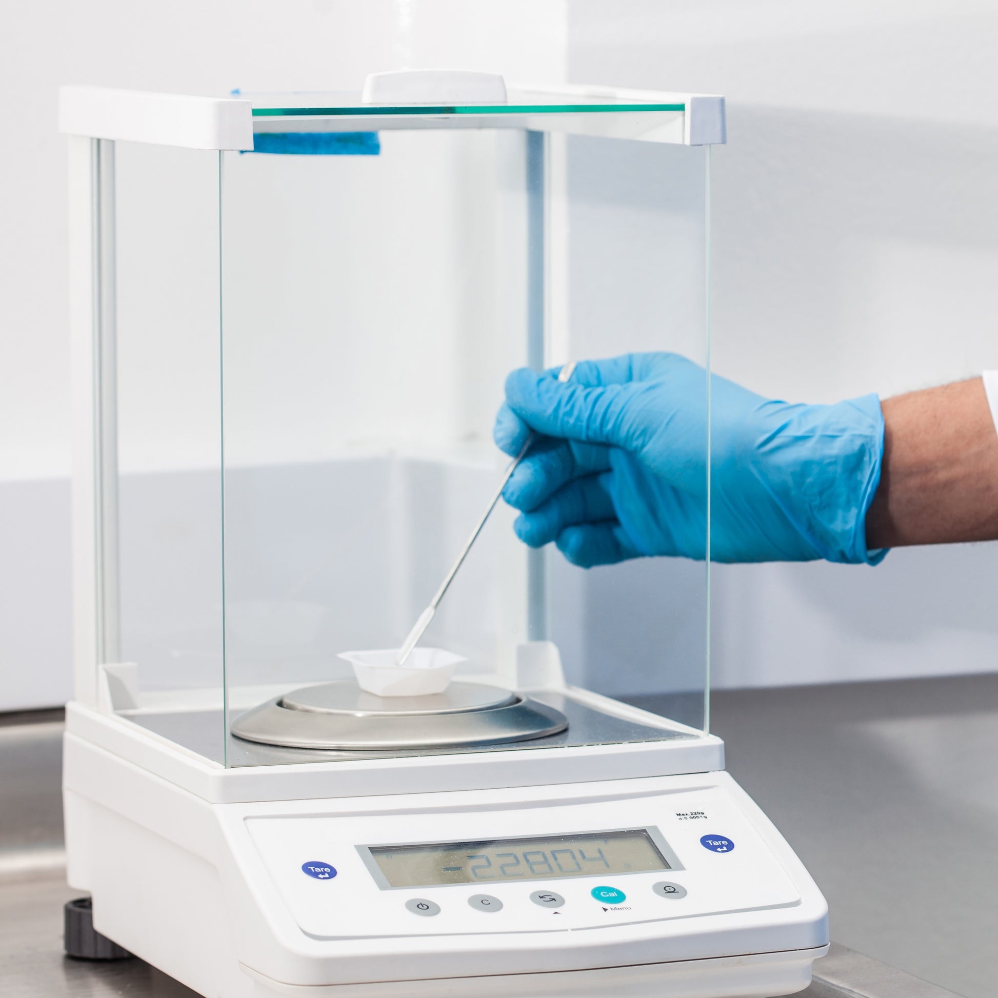 <Male hand with blue glove adding powder with a spatula to a weigh boat on a white, digital analytical balance with clear glass screens. Credit> AdobeStock_178852086