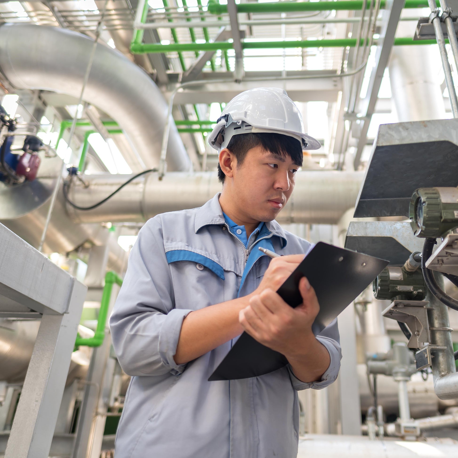 <Male engineer, wearing grey overalls and white hard hat, stood in the production area surrounded by stainless steel pipework taking readings from a meter on a clipboard. Credit> AdobeStock_196127416