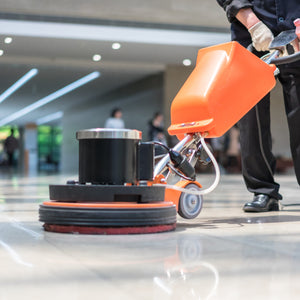 <Legs of a man dressed in black uniform using a floor polisher on the cream marble floor of a floodlit office reception with employees blurred in the background entering the building. Credit> AdobeStock_205886837