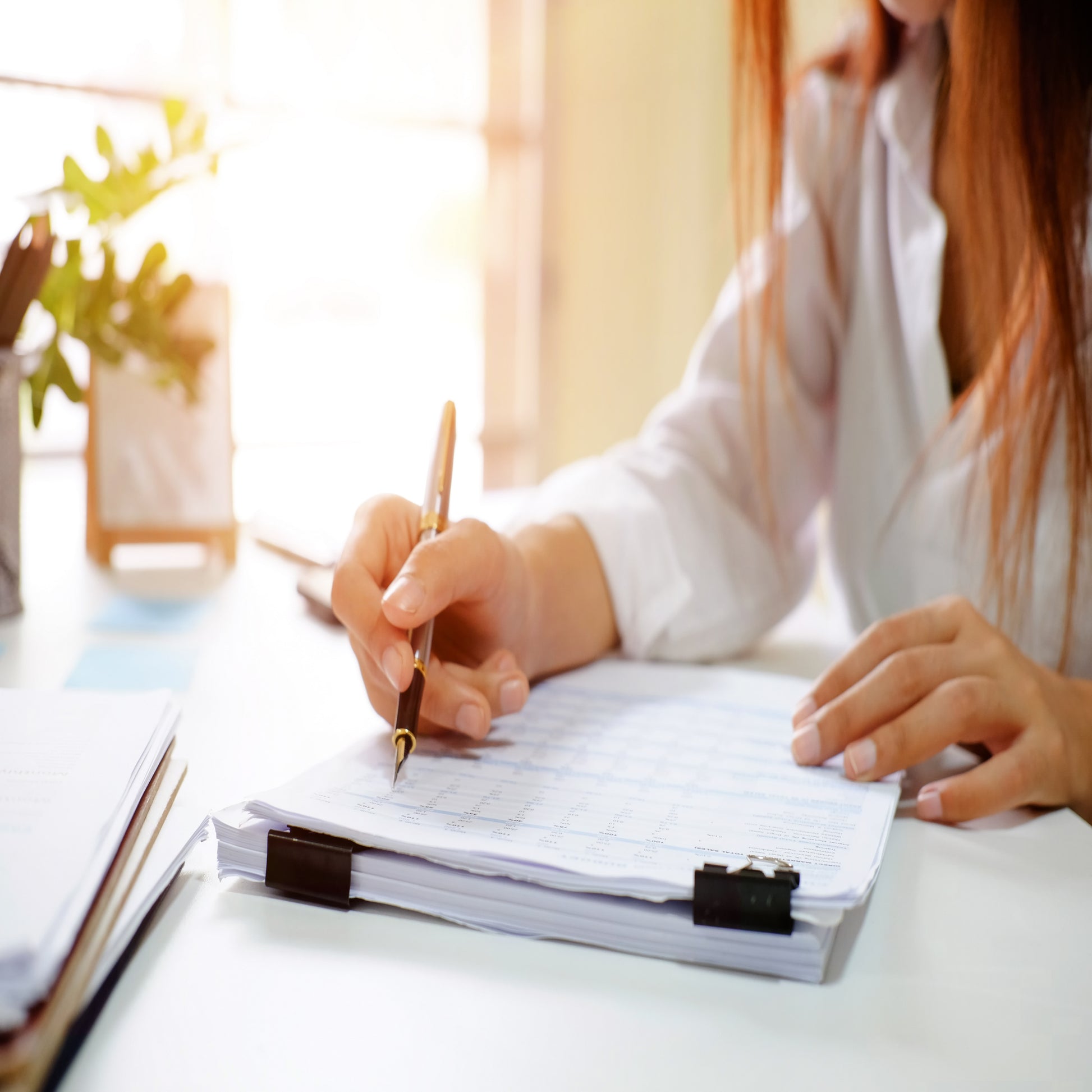 <Arms and torso of a brunette woman, wearing a white shirt, holding a gold fountain pen, which she is using to review data on two piles of papers held together with binder clips. Credit> AdobeStock_208566614