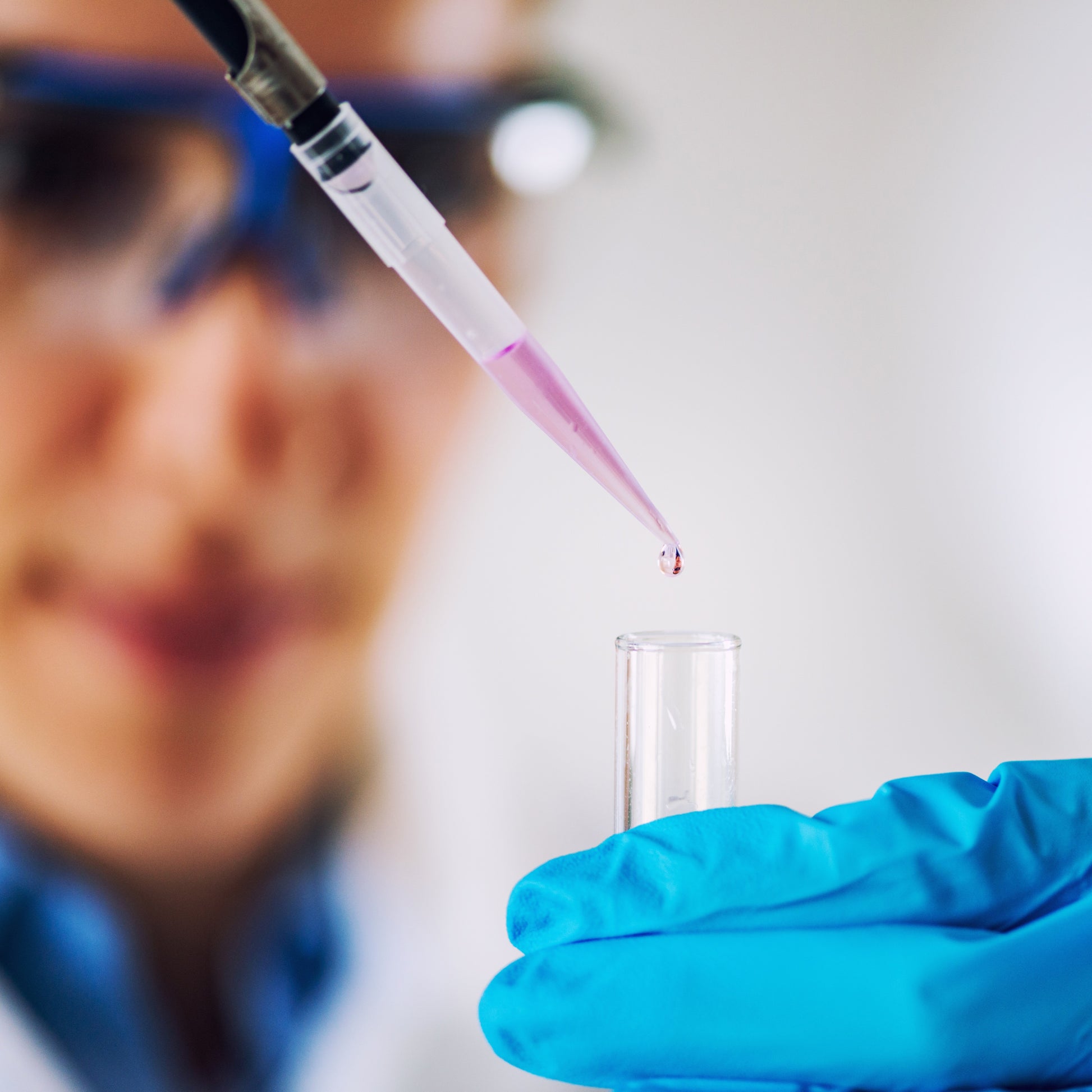 <Blurred laboratory analyst wearing safety specs in background with close up of pipetting a clear, purple liquid into a test tube. Credit> AdobeStock_212916043