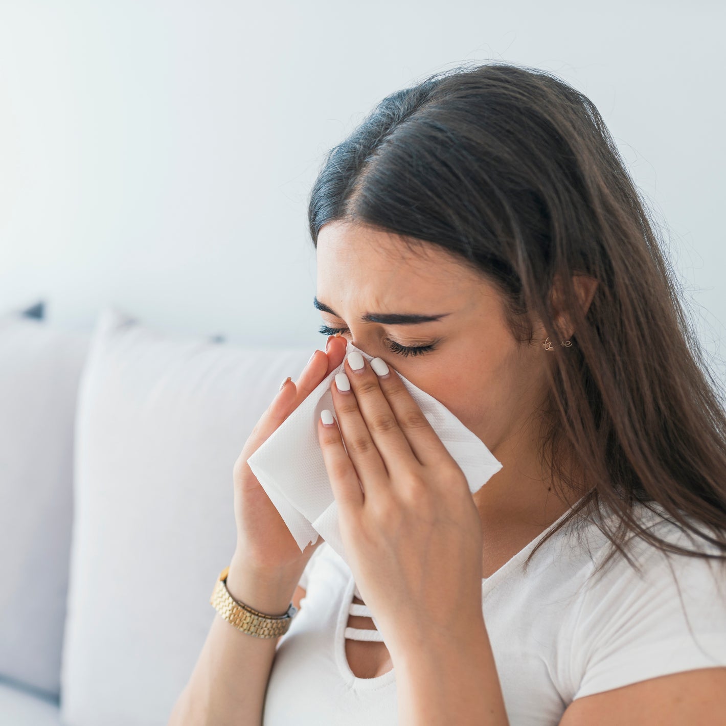 <Brunette female employee, wearing a white t-shirt and gold watch, off work sick sitting on a white sofa at home blowing her nose into a tissue with cold and flu symptoms. Credit> AdobeStock_219422085