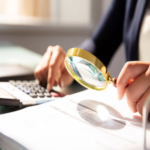 <Arms of a woman holding a bright, gold magnifying glass in her left hand to review a document whilst typing on a laptop keyboard with her right hand. Credit> AdobeStock_224910698