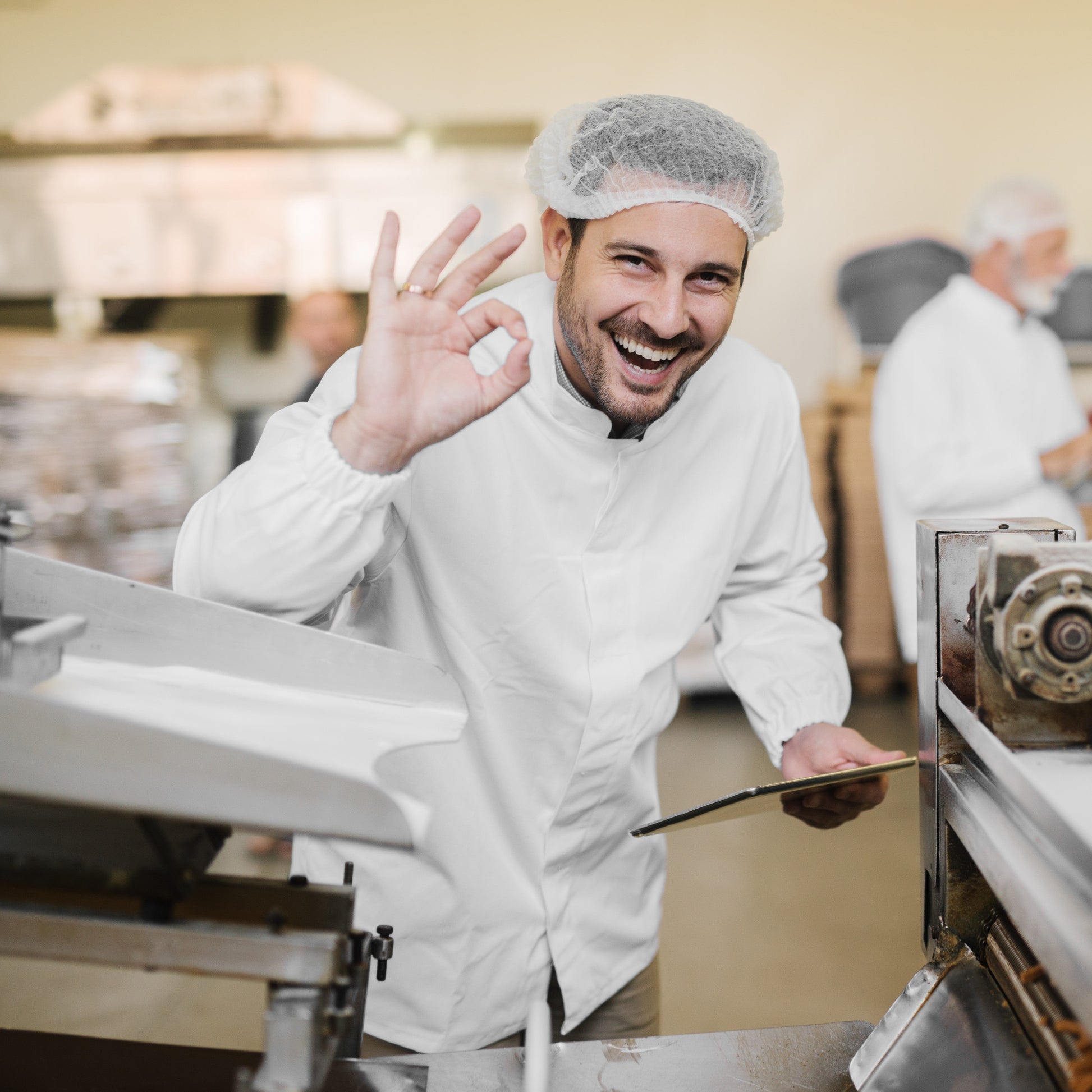 <Male, production team leader, wearing white coat and hairnet, smiling and making an OK symbol with his hand next to a tablet filling machine. Credit> AdobeStock_226047743