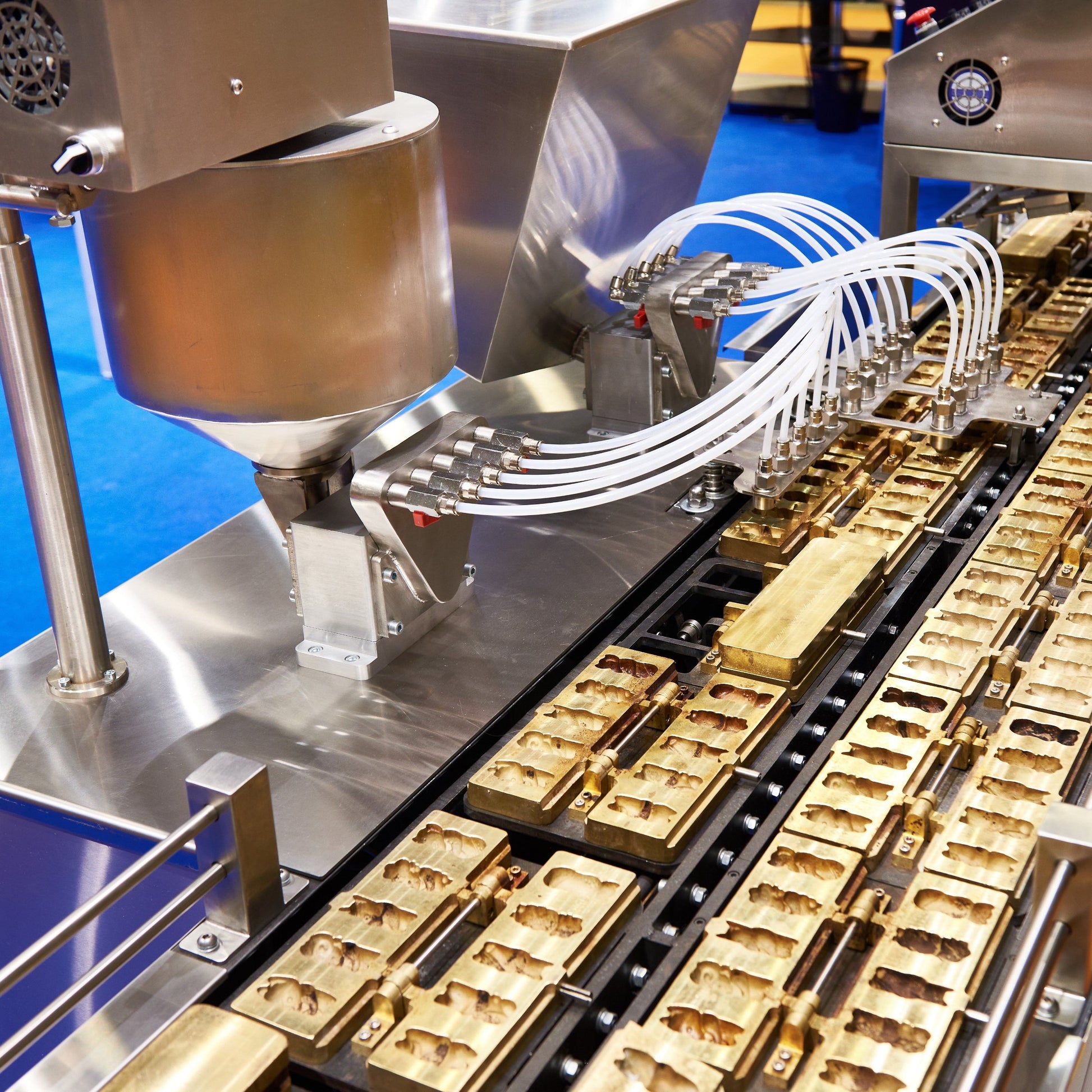 <Series of 5 plastic tubes coming from a stainless steel hopper filling gold coloured metal lozenge moulds with hot molten dietary supplement confectionery and passing them down a conveyor into a cooling tunnel with a blue non slip floor. Credit> AdobeStock_227896938