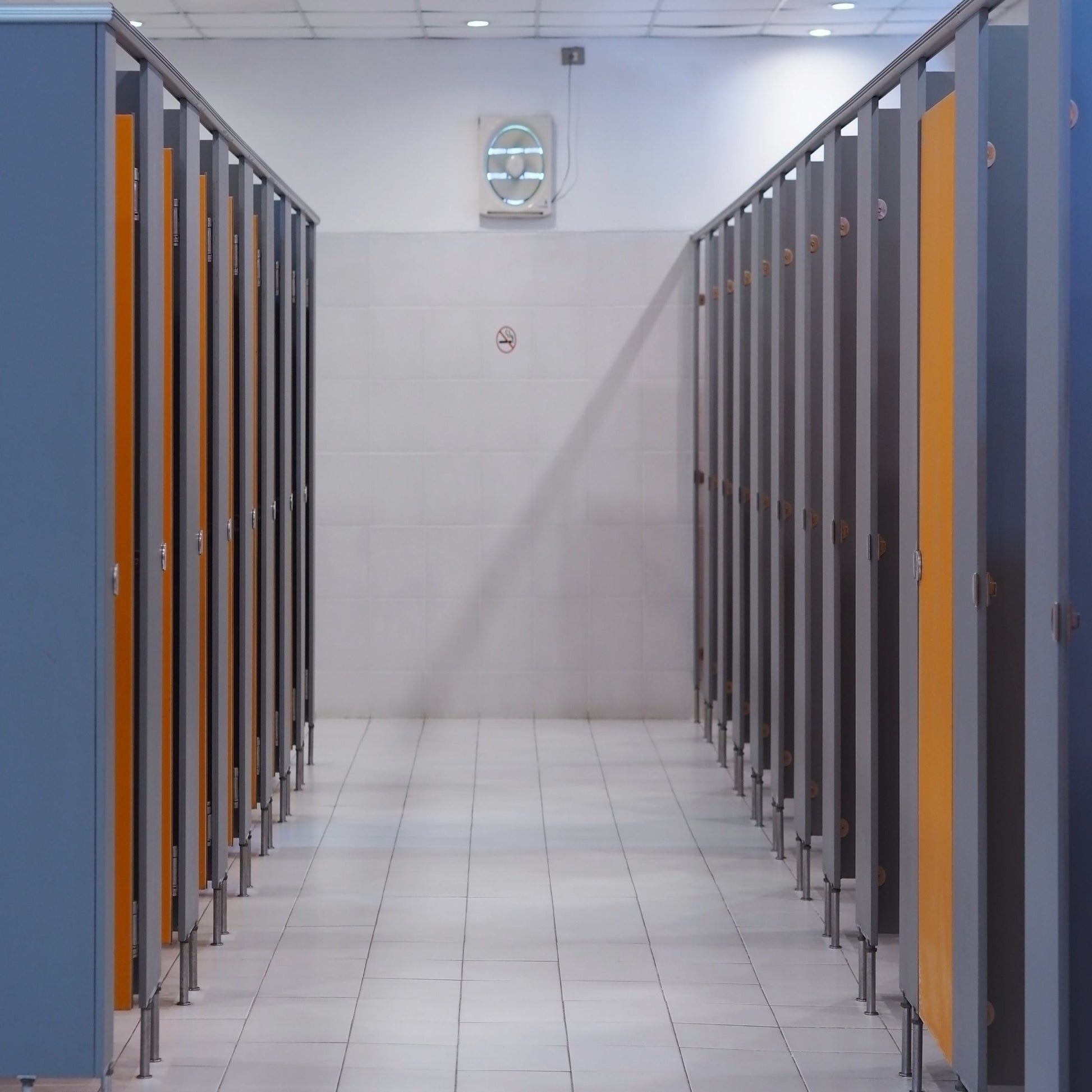 <Employee series of blue grey toilet cubicles with wooden doors in a rest area with grey walls, white tiled floors and white ceiling grid with wall ventilation fan. Credit> AdobeStock_239768281