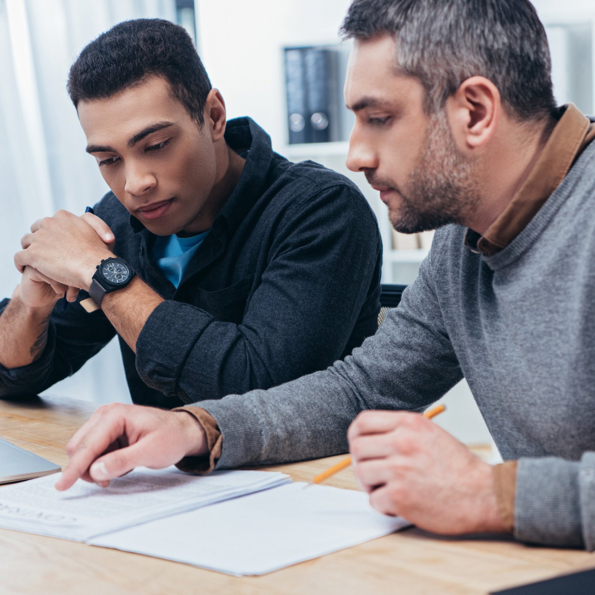 <Two male supervisors, wearing navy blue and grey jumpers,  sat at a wooden office desk reviewing production paperwork, one of them pointing at something in the documents. Credit> AdobeStock_241546209