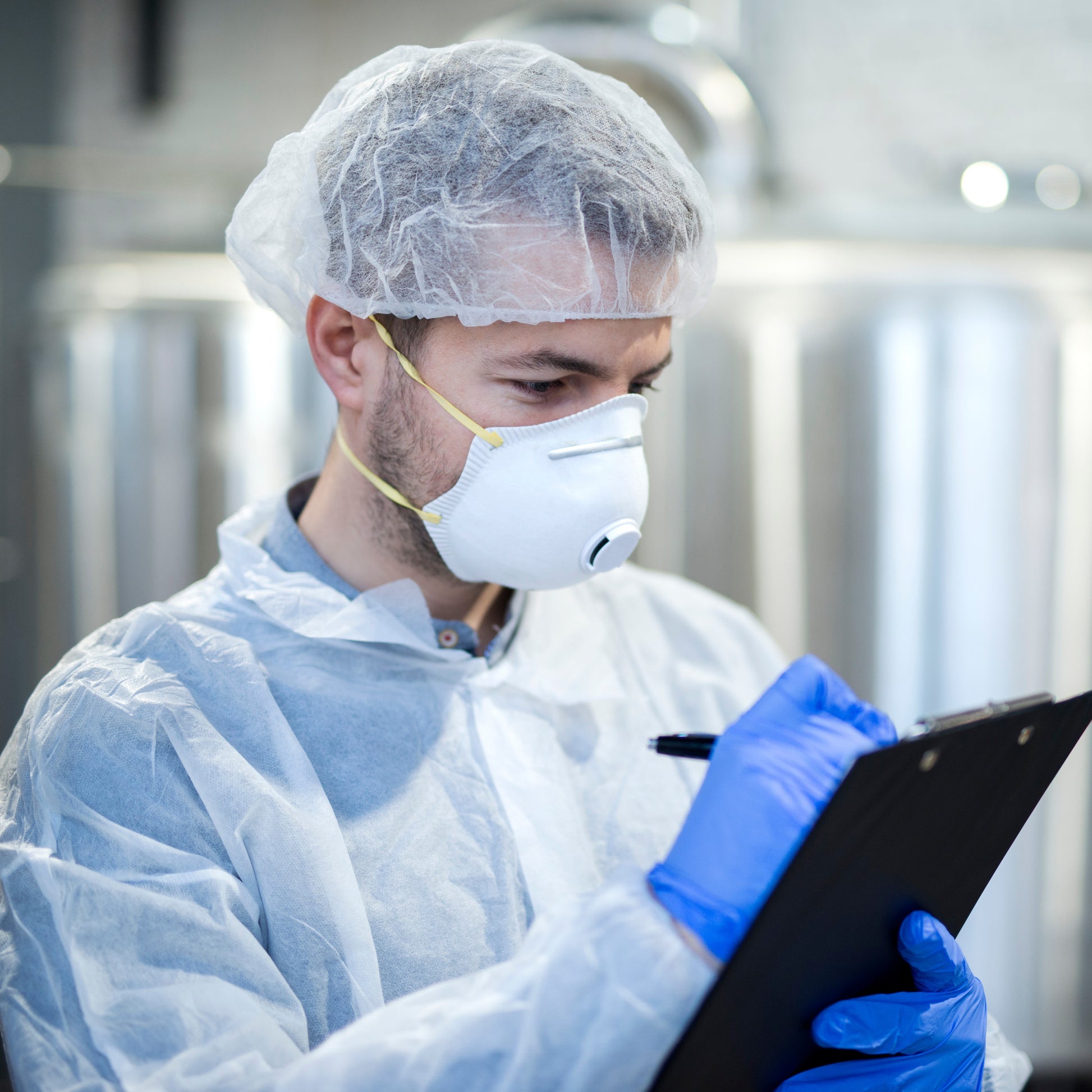 <Male, bearded production manager wearing a white coat, hairnet, face mask and blue gloves completing a clipboard checklist with a black pen. Credit> AdobeStock_244373126