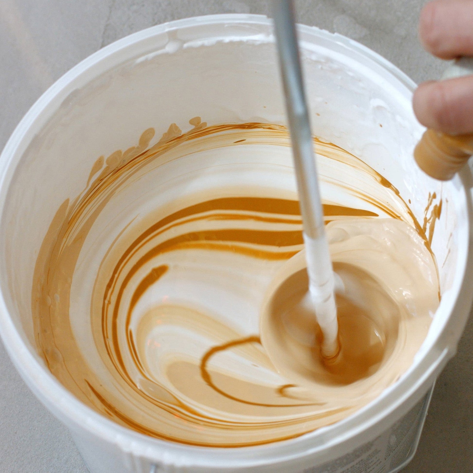 <Hand of a manufacturing operator adding a brown liquid from a bottle to a white cream in a white bucket with an air propellor mixer causing a vortex and swirls of colour. Credit> AdobeStock_269910716