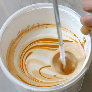 <Hand of a manufacturing operator adding a brown liquid from a bottle to a white cream in a white bucket with an air propellor mixer causing a vortex and swirls of colour. Credit> AdobeStock_269910716