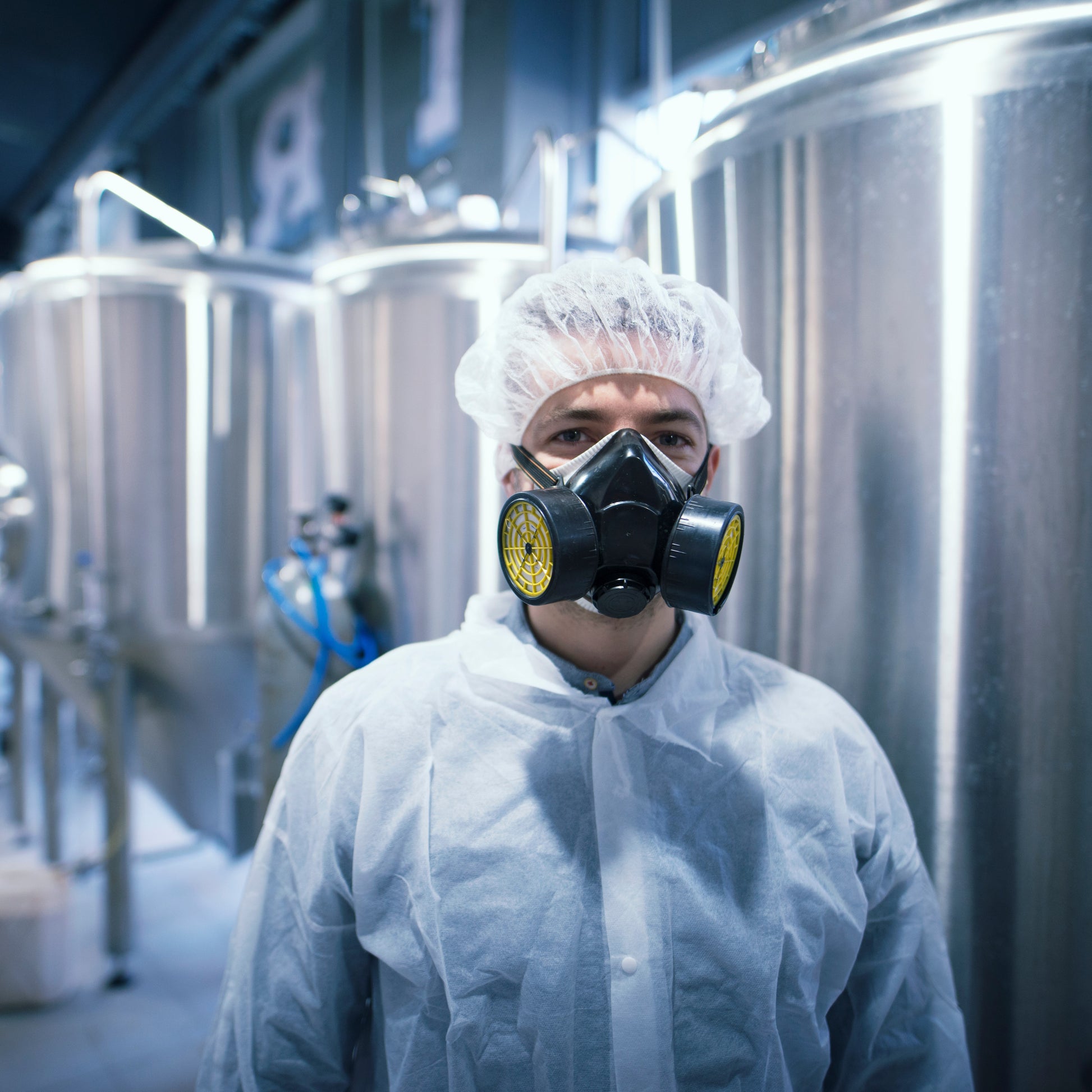 <White male production operator stood in a manufacturing area of mixing tanks wearing a white coat and hairnet with a protective dust mask covering his nose and mouth. Credit> AdobeStock_271852211