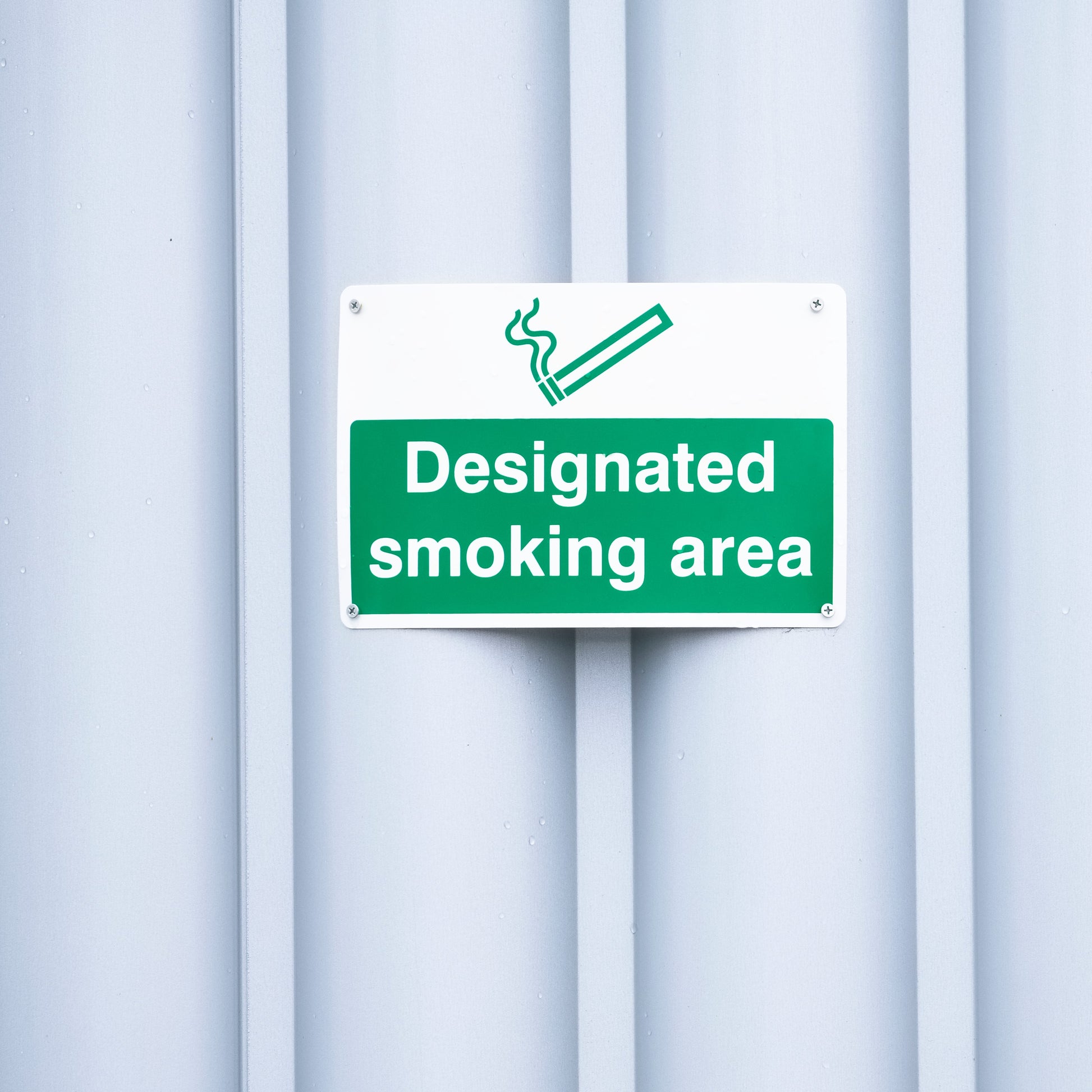 <Green sign on a white corrugated metal building to direct personnel to a designated smoking area on site. Credit> AdobeStock_272303046