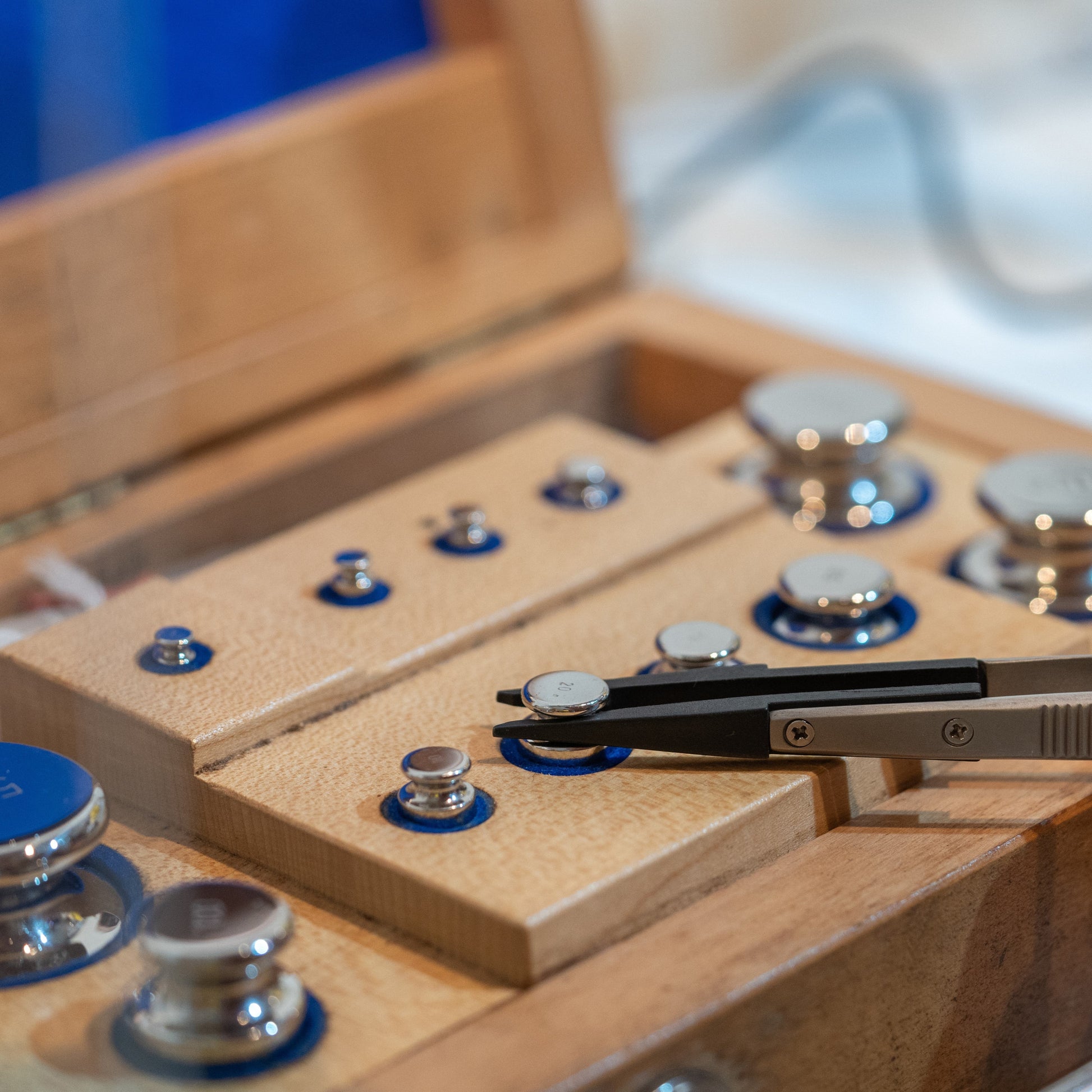 <Tweezers picking up a chrome calibration weight for an analytical balance from a wooden box set of standard weights. Credit> AdobeStock_274078427