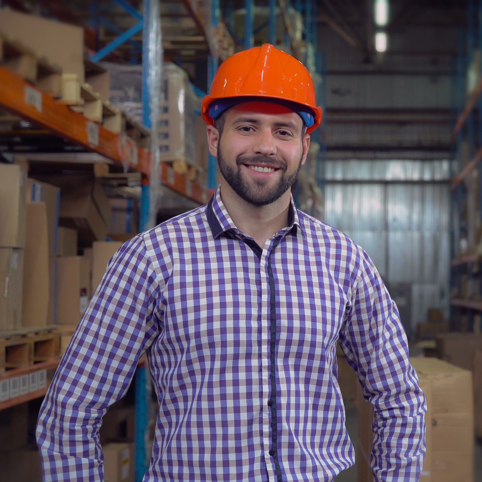 <Experienced and confident bearded male warehouse manager wearing a blue checked shirt and orange safety hard hat stood in front of racking containing pallets of boxes. Credit> AdobeStock_277436381