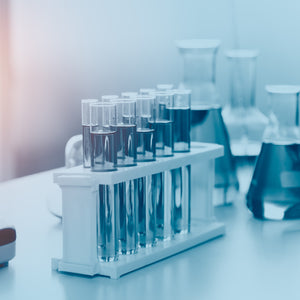 <Under ultra violet filter, white test tube rack on a white surface with eight test tubes and three large flasks next to it all containing violet blue liquid. Credit> AdobeStock_278678145