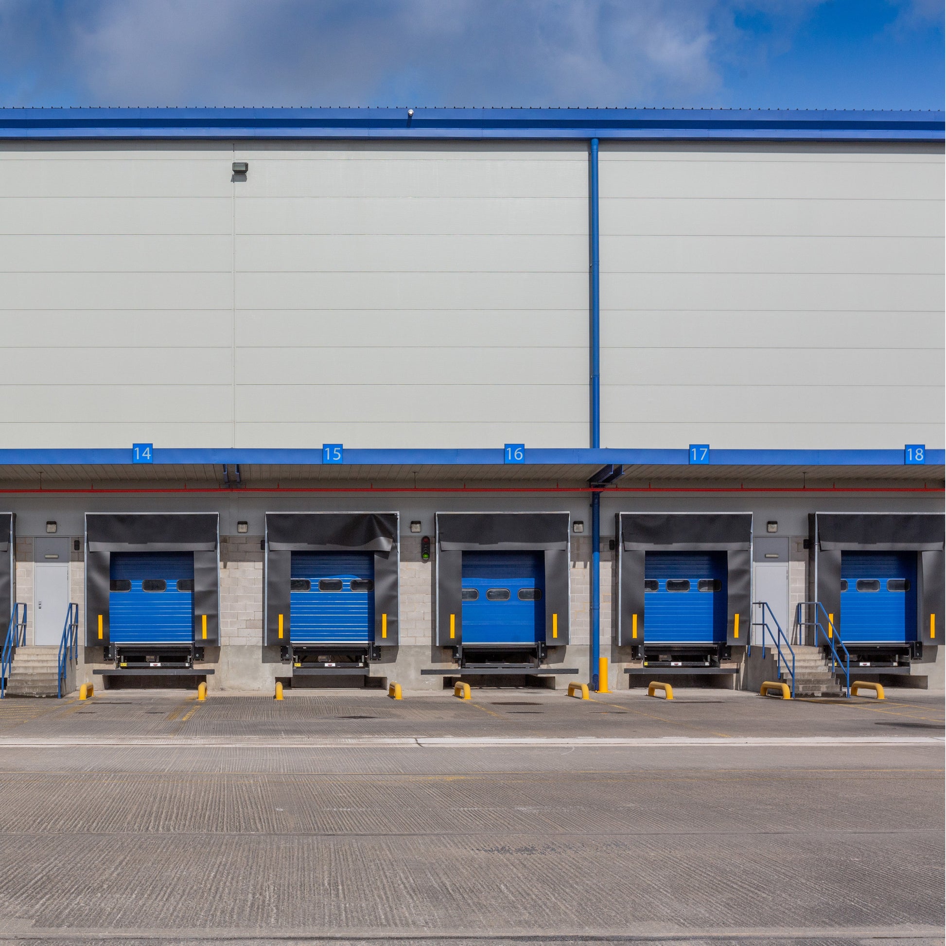 <View from the outside concrete yard in sunshine of five blue roller shutter dock doors for loading and unloading lorries into a grey metal building numbered 14 to 18. Credit> AdobeStock_292350584