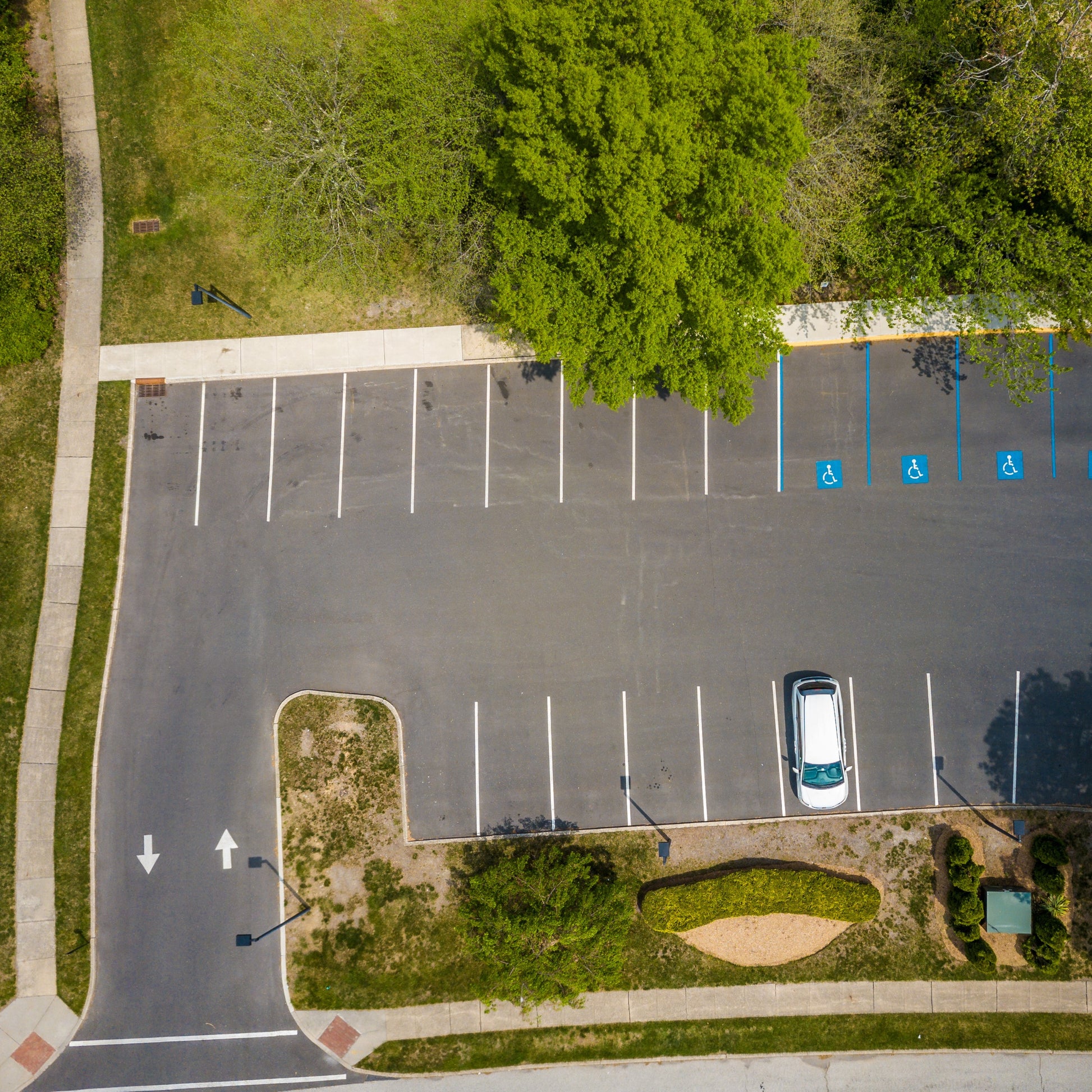 <Employee tarmac car park or parking lot of 20 spaces and 4 disabled driver spaces with one white car surrounded by grass, trees and pathways. Credit> AdobeStock_294267869