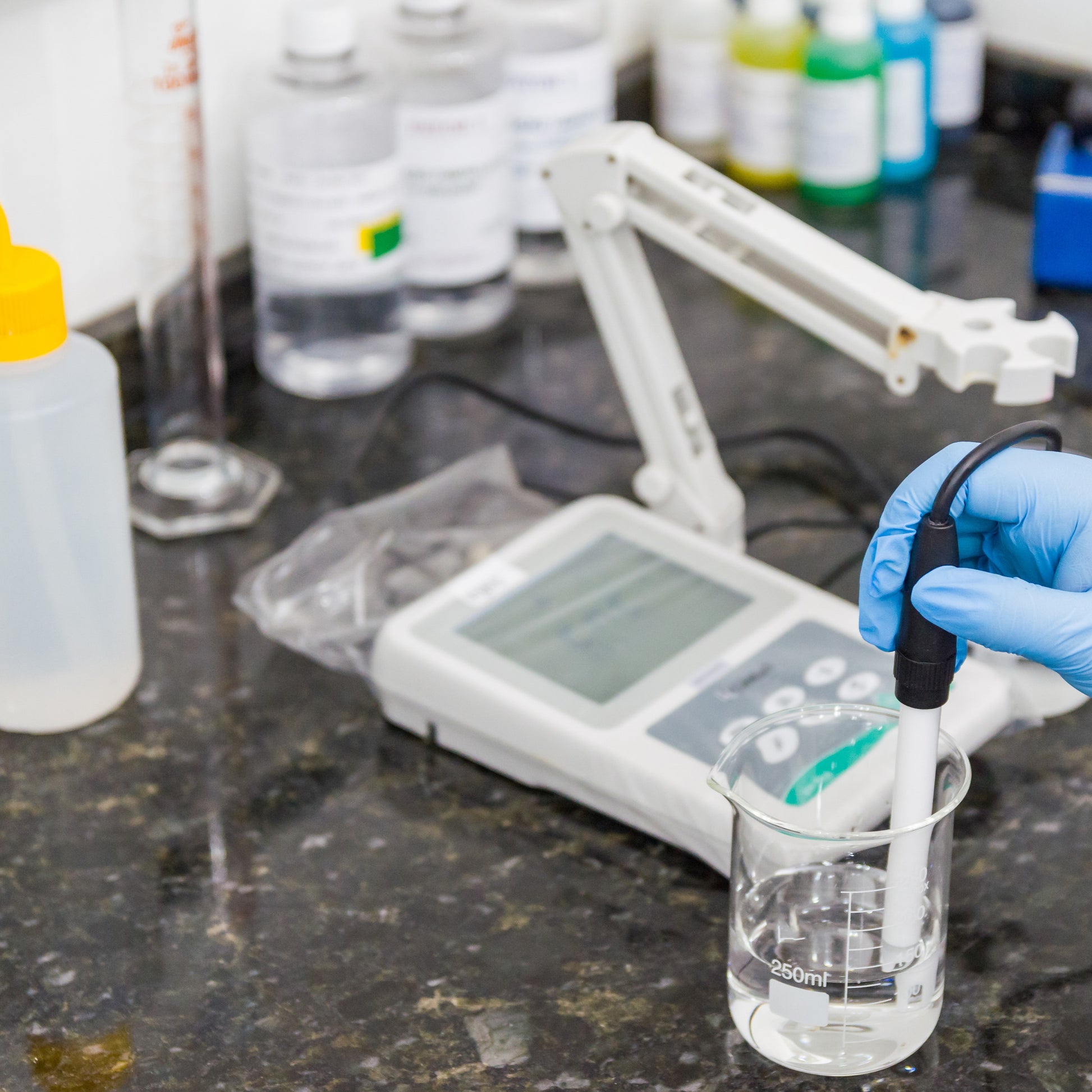 <Hand in a blue glove holding the probe of a pH meter in a glass beaker of clear liquid, on a stone effect worktop, with bottles of buffer solution. Credit> AdobeStock_296409151