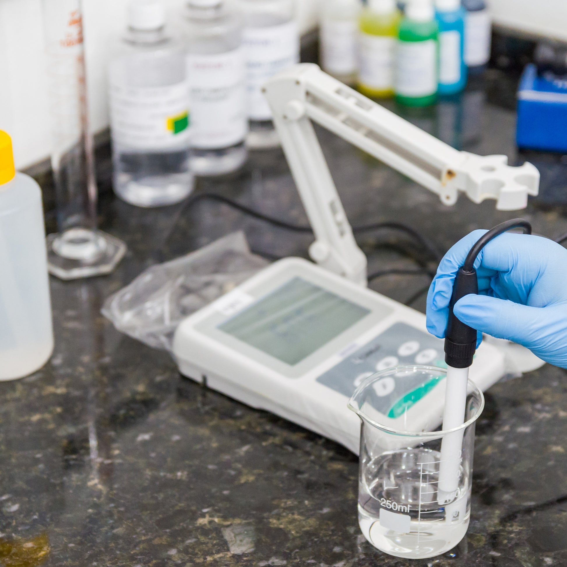 <Hand in blue glove holding a white and black probe in a beaker of clear liquid attached to a combined Conductivity and pH meter. Credit> AdobeStock_296409151