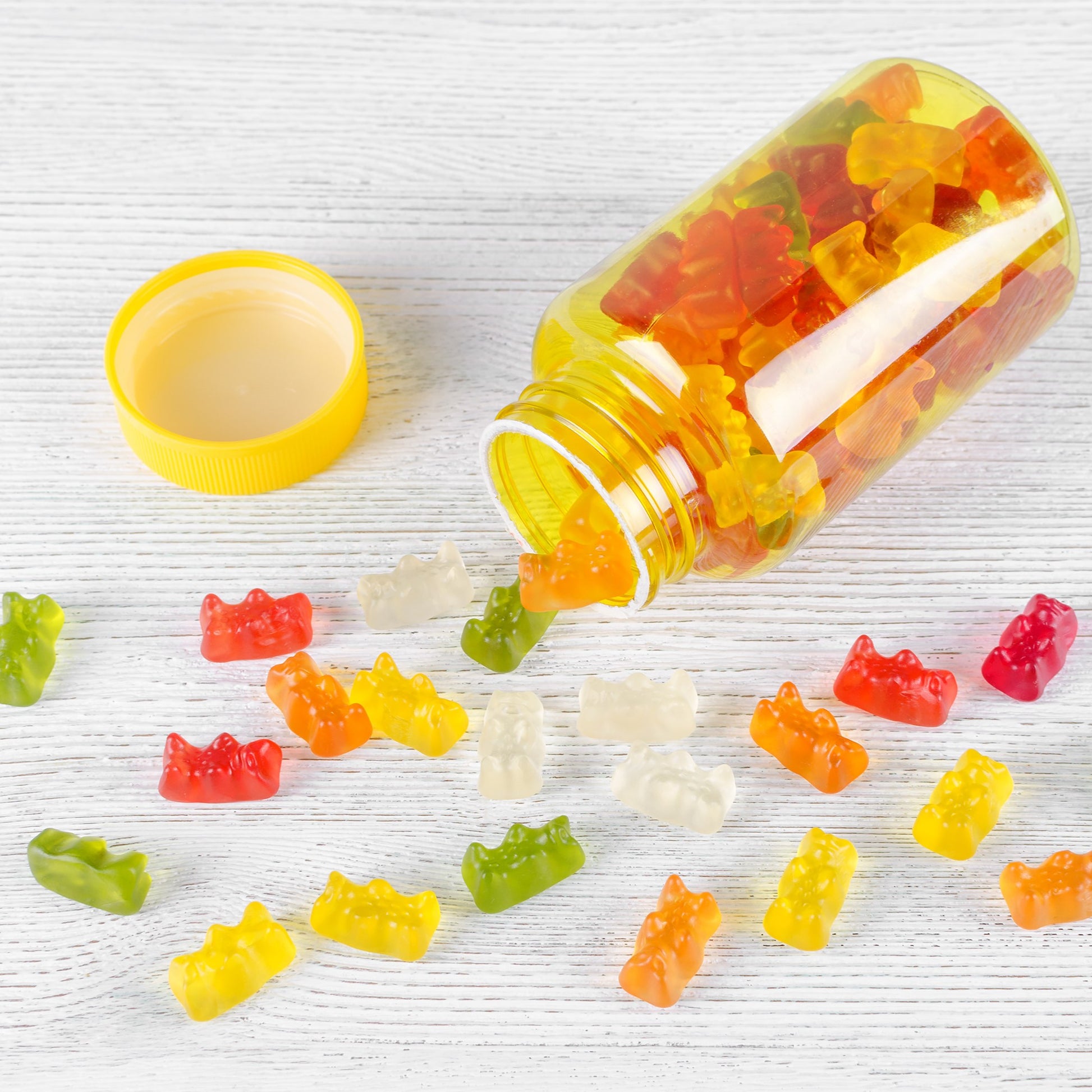 <Clear, yellow plastic bottle, with opaque yellow lid, on its side with 15 red, orange, yellow, and green gummy bears tipped out on to a white woodgrain effect table. Credit>  AdobeStock_301147051 