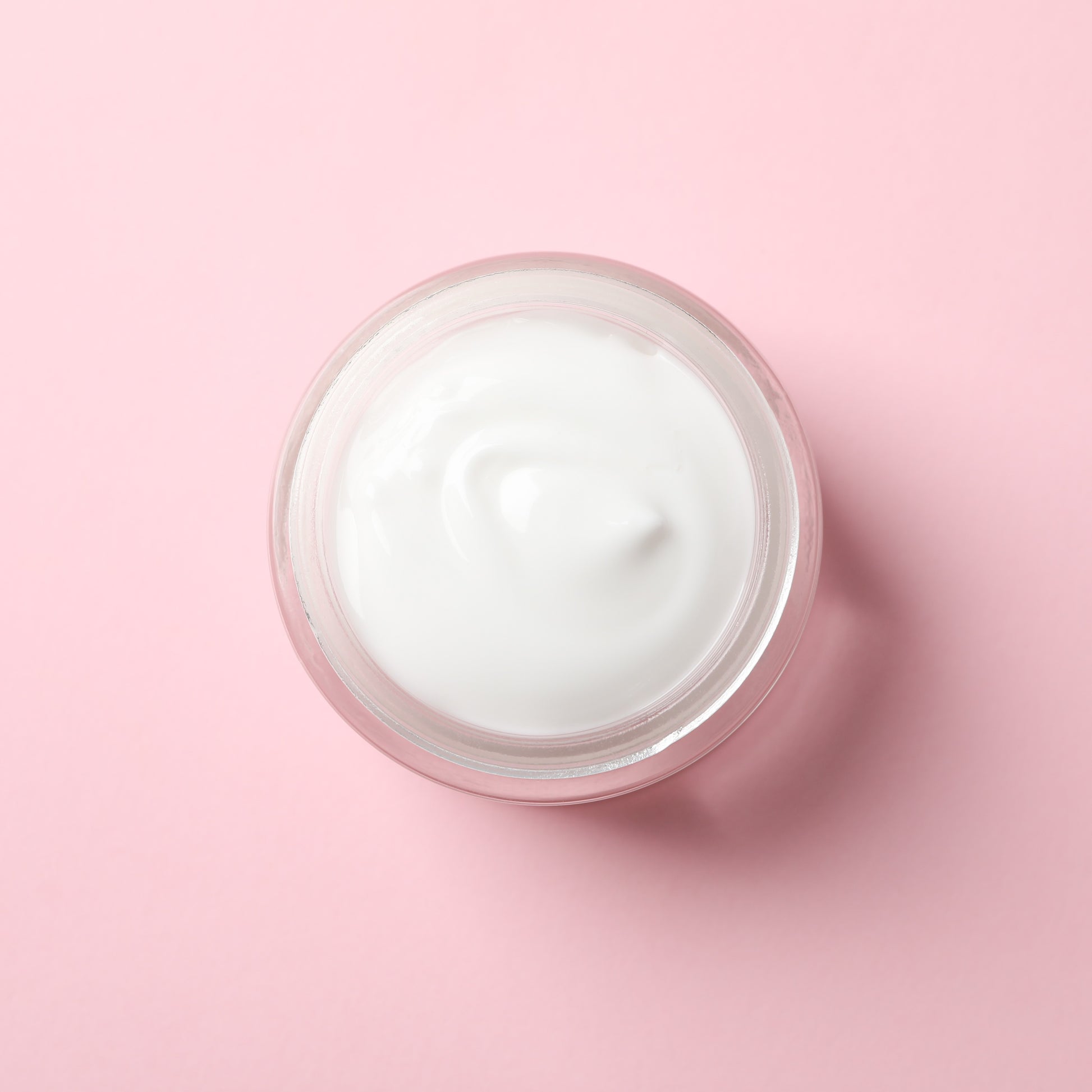 <Overhead view of a viscous hand cream filled into a round, clear plastic jar sitting on a bright light pink surface. The cream seems to be at a viscosity that has allowed it to spread and fill the jar evenly but alos maintain a peak shape once it stopped filling. Credit> AdobeStock_305417987