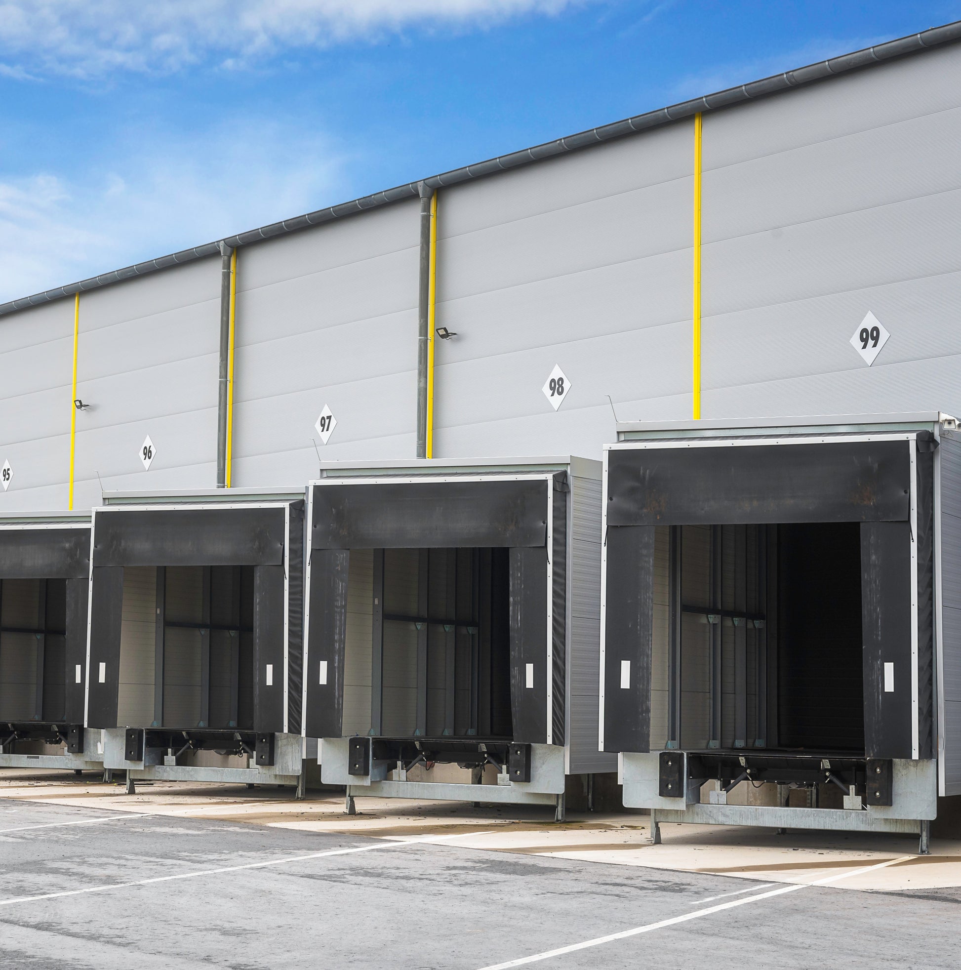 <From the concrete yard for distribution, four loading bay doors in the side of a grey metal warehouse at lorry height and shape numbered 95 to 99. Credit> AdobeStock_326508468