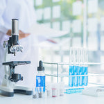 <White, black, and chrome microscope on a  white lab surface next to a rack with 4 test tubes of blue liquid and a lab analyst in white labcoat in a blurred background with  windows and green outside area. Credit> AdobeStock_336473215