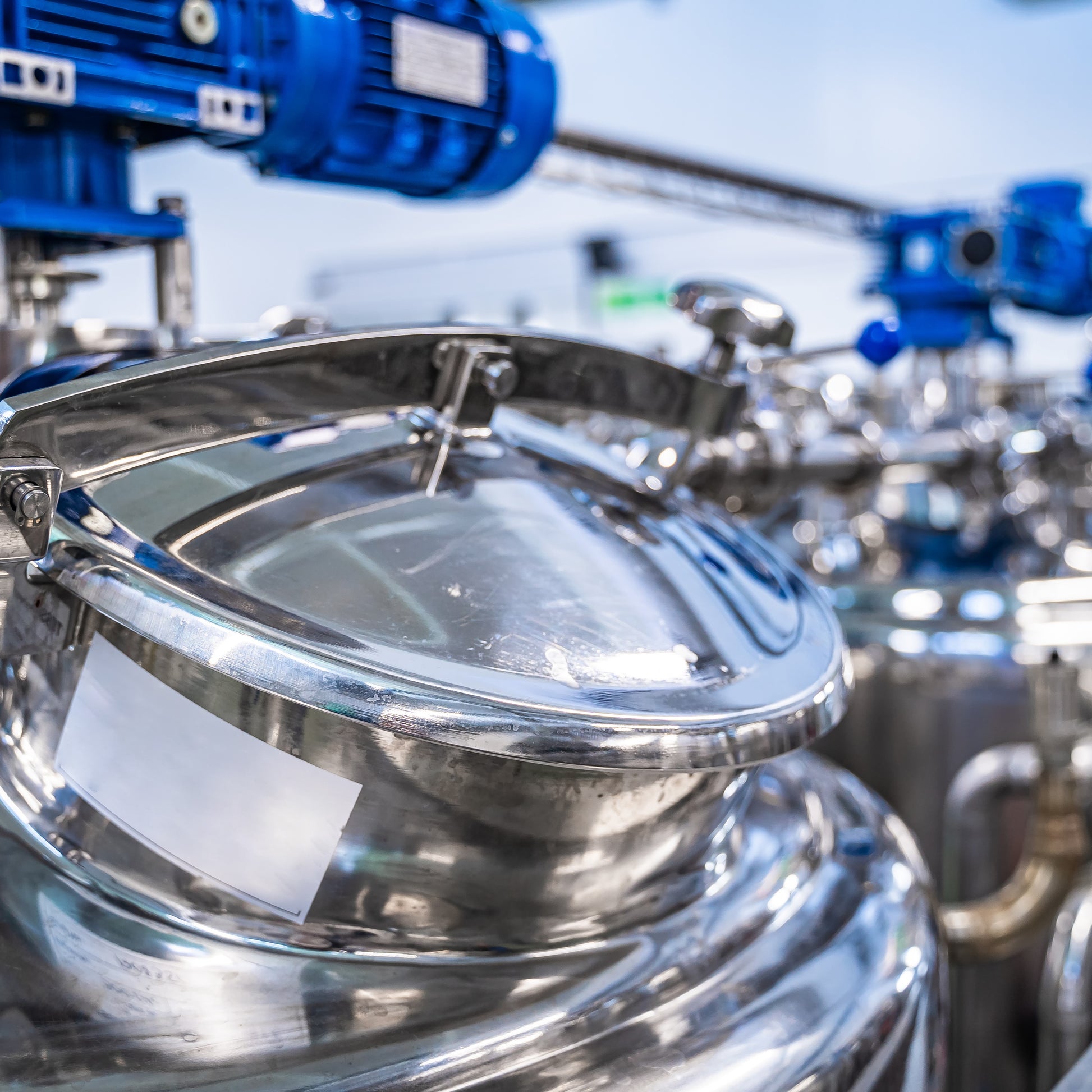 <Entry port or lid of a polished, mirror finish, stainless steel mixing vessel or tank, with an overhead blue mixer or homogenizer with another in the background. Credit> AdobeStock_336493389