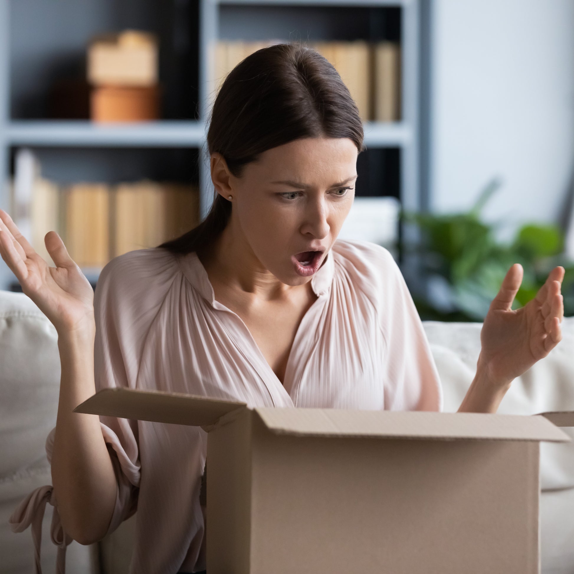 <Female consumer wearing cream coloured top, sitting on white sofa in living room, shelving and plant behind her, visibly shocked with hands in the air and mouth open at the box she has opened from an online pharmacy. Credit> AdobeStock_345250156