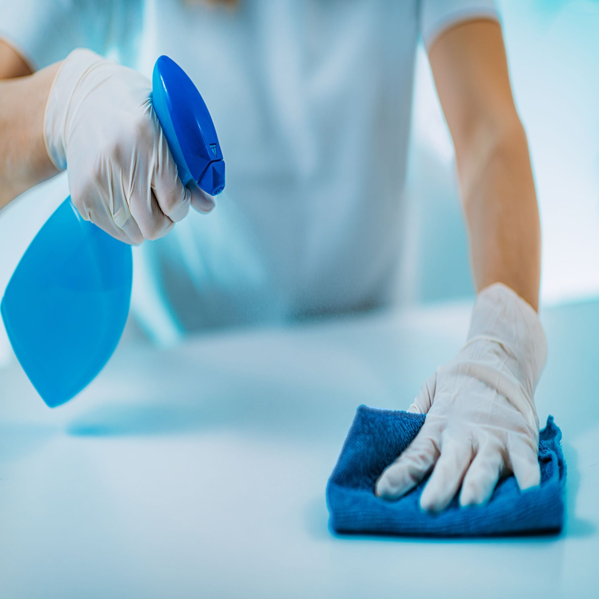 <Arms and body of a woman wearing light blue overall and white gloves cleaning a white contact surface with a clean blue cloth and an approved  cleaning spray from a blue spray gun. Credit> AdobeStock_346099456