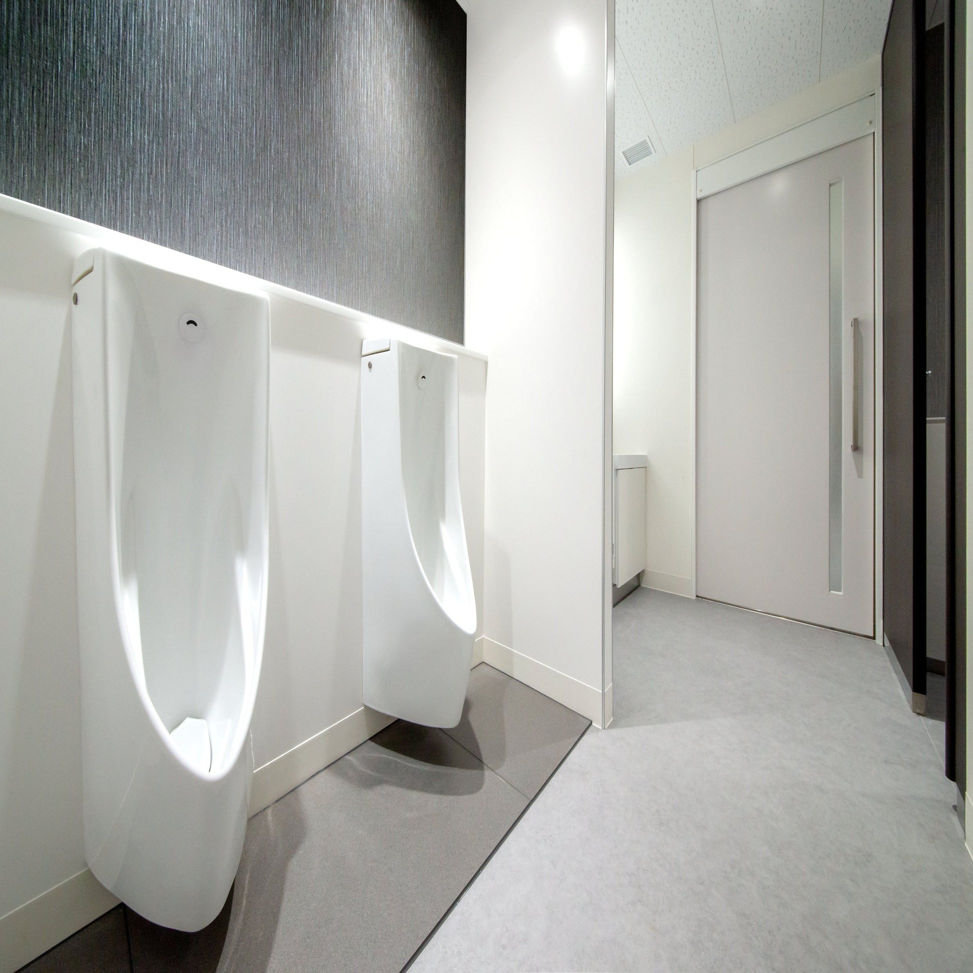 <Two white urinals in an employee rest area with dark grey stone effect wall, grey vinyl floor and white walls and doors. Credit> AdobeStock_364201756