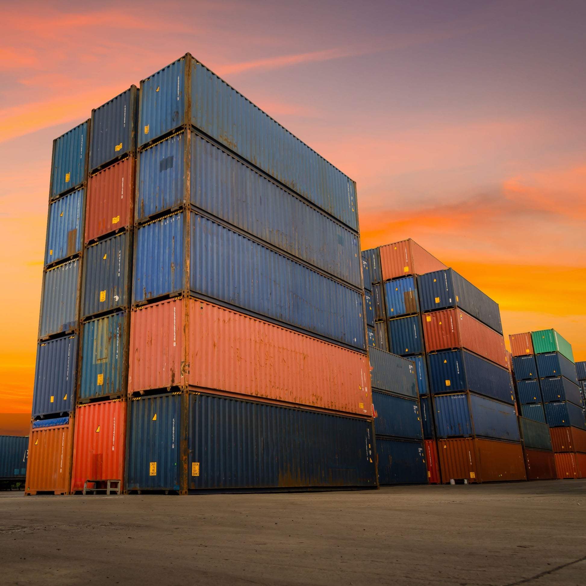 <Stack of blue and orange shipping containers five high at a concrete port in winter sunset ready to be distributed by sea. Credit> AdobeStock_377498722