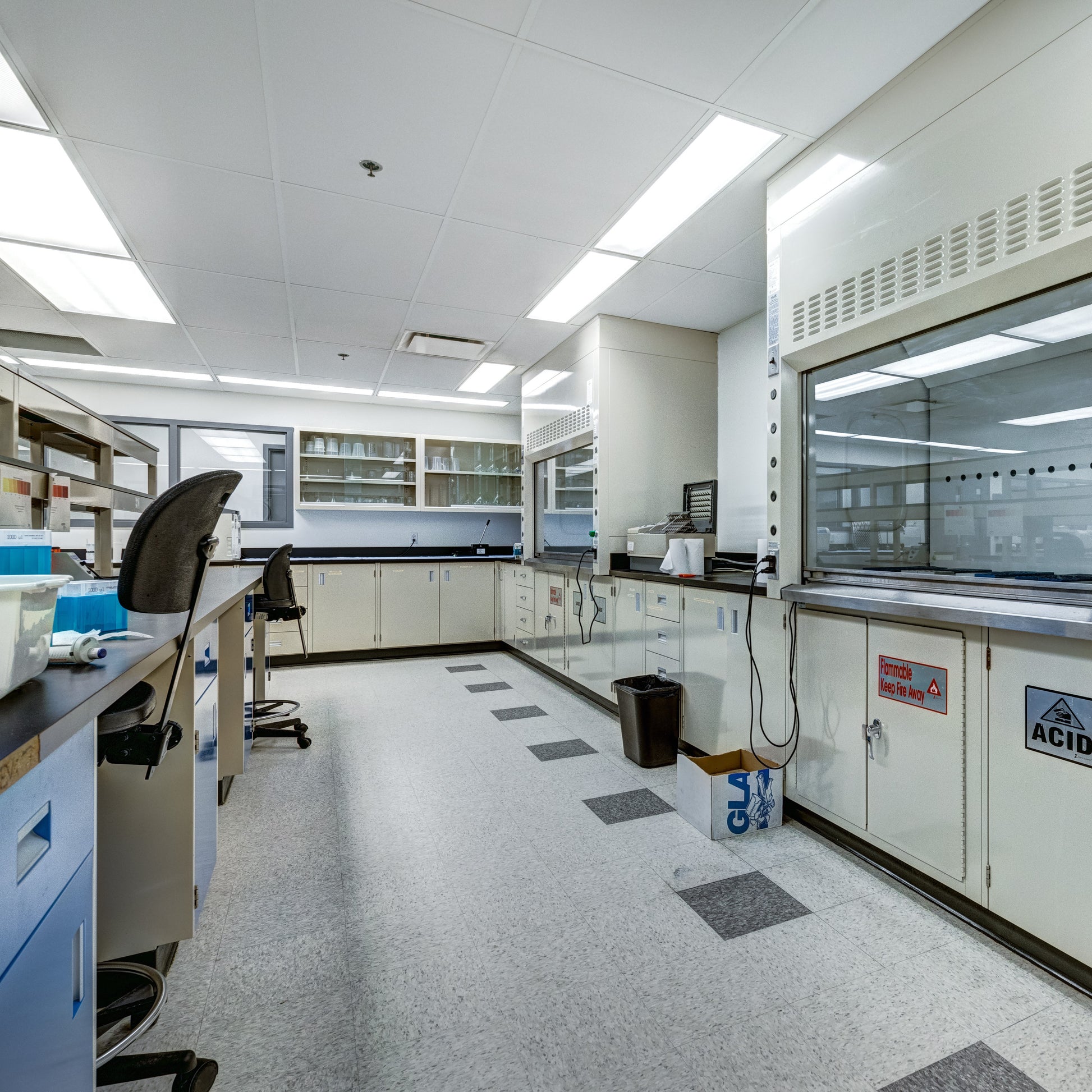 <Laboratory with cream cupboards on one side, blue on the other, grey non-slip floor, white suspended ceiling with square LED lights, a fume cupboard with hood. Credit> AdobeStock_378631743