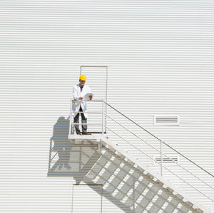 <Man wearing white coat and yellow hard hat stood outside a door in sunshine in the middle of a white corrugated metal face of a building at the top of a stairwell. Credit> AdobeStock_383183290