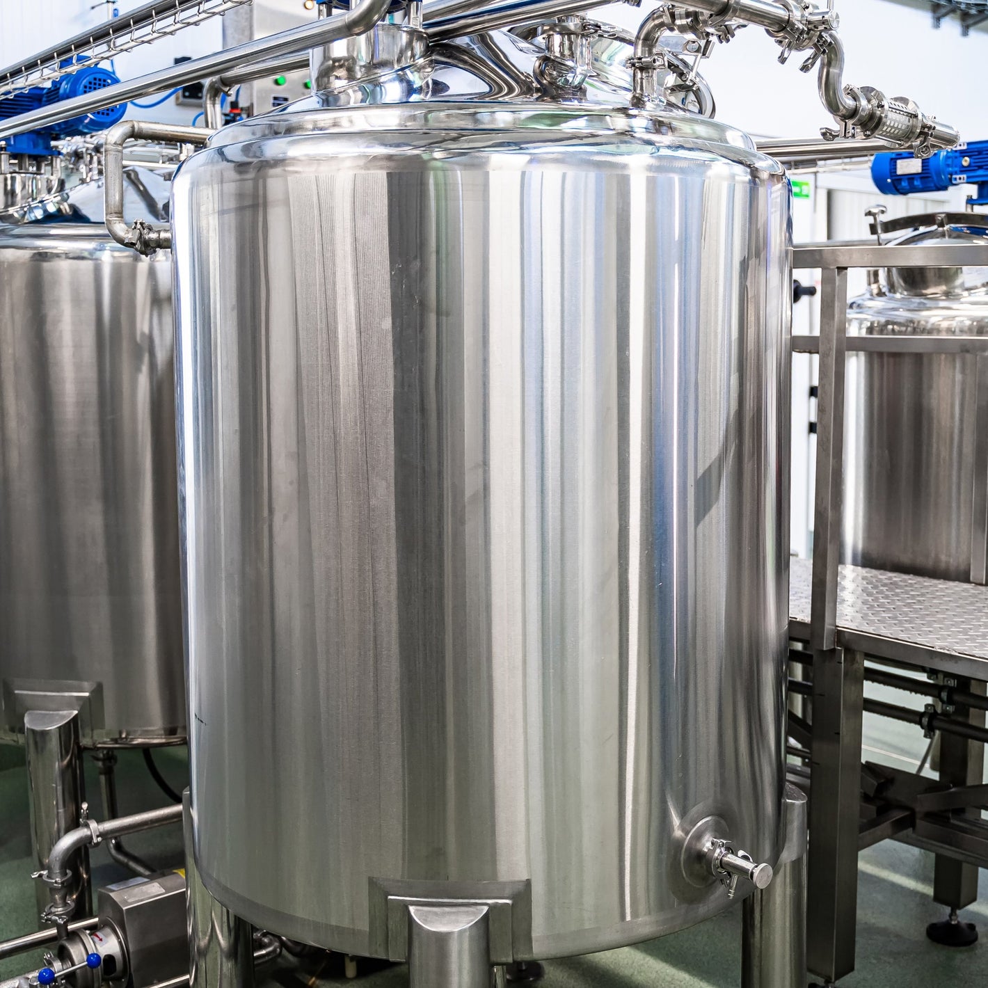 <Several large, cylindrical, shiny stainless steel  mixing tanks and pipework in a dietary supplement manufacturing unit with gantry, grey non slip floor and white ceiling. Credit> AdobeStock_383238064