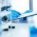 <Hands of a laboratory analyst wearing blue gloves holding a pen and completing a phase 1 OOS investigation on a clipboard in blurred lab. Credit> AdobeStock_391585993