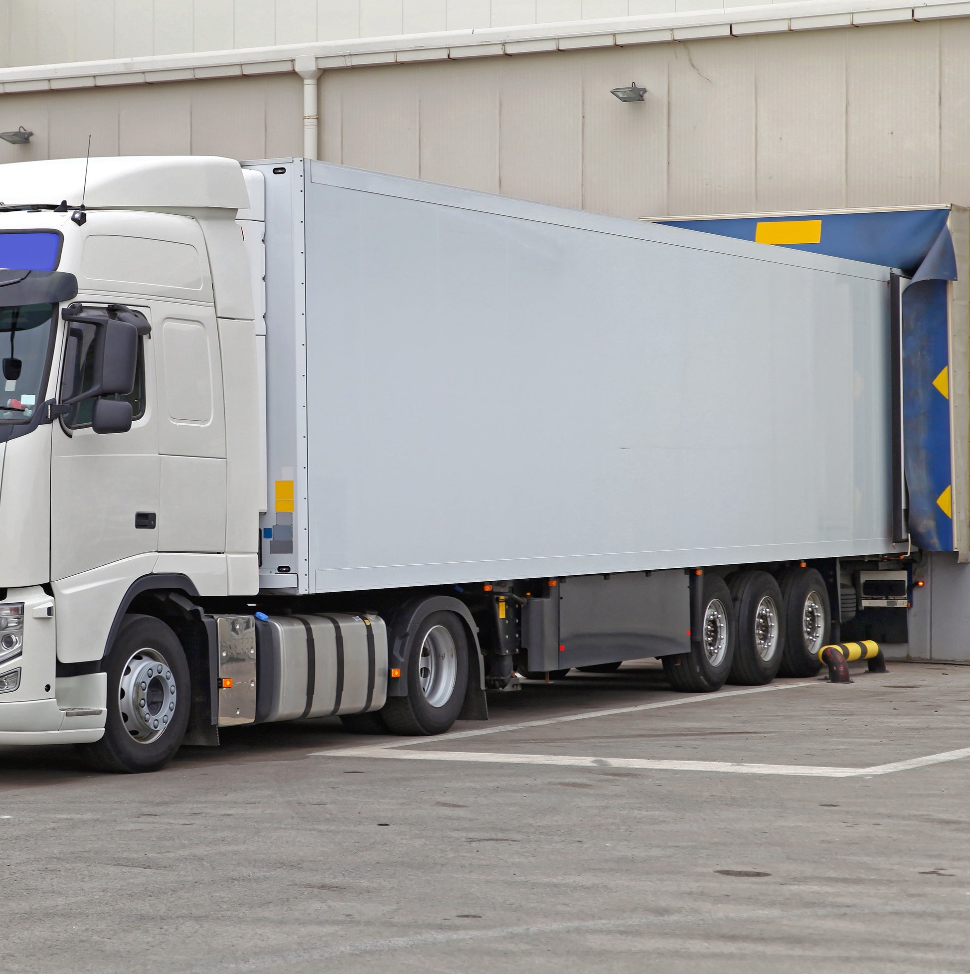 <Large white lorry in the concrete yard of a drug facility reversed up to a dock door in the warehouse to make a delivery. Credit> AdobeStock_403412047