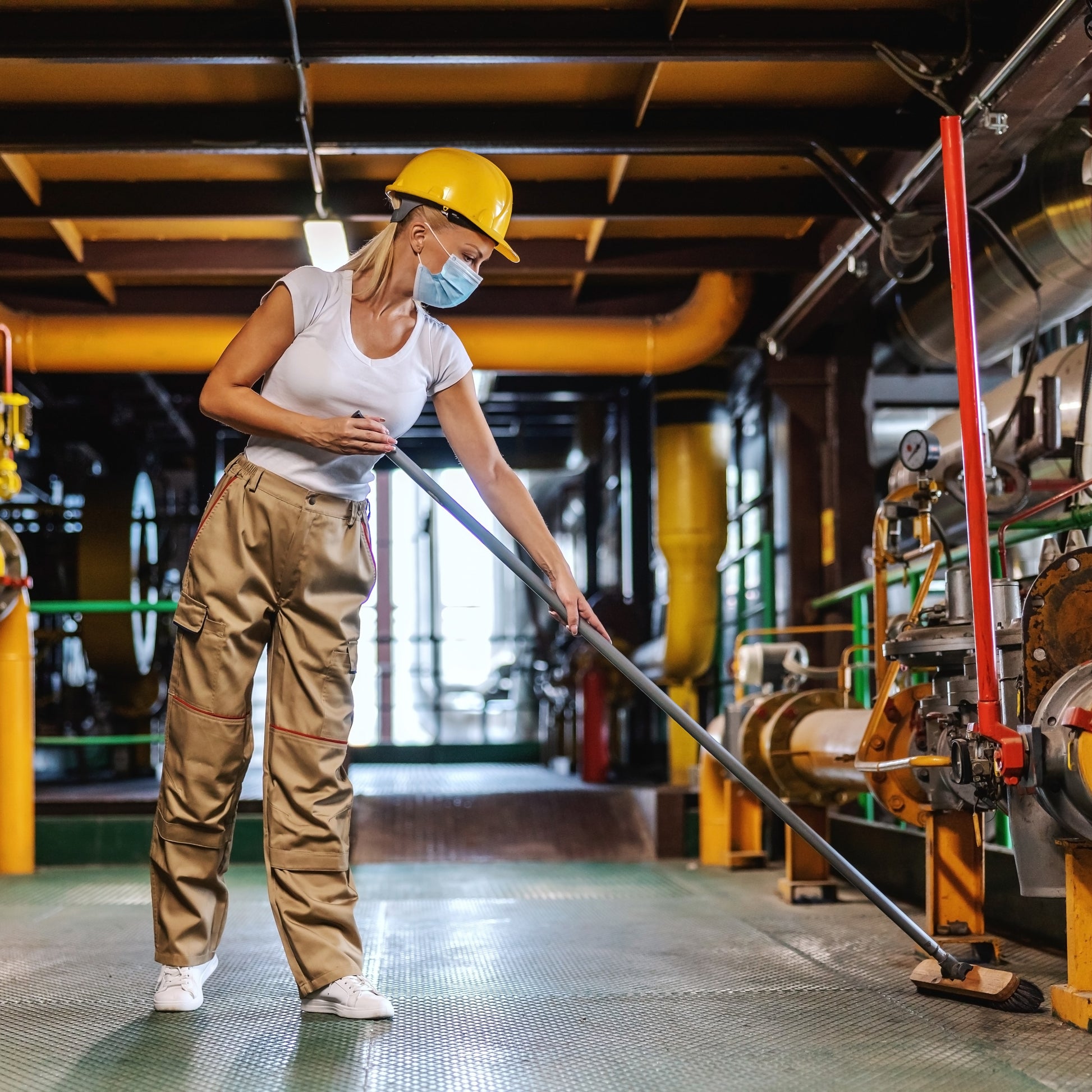 <Female production operator cleaning the metal grid floor of a plant room area with a brush. Credit> AdobeStock_403490907