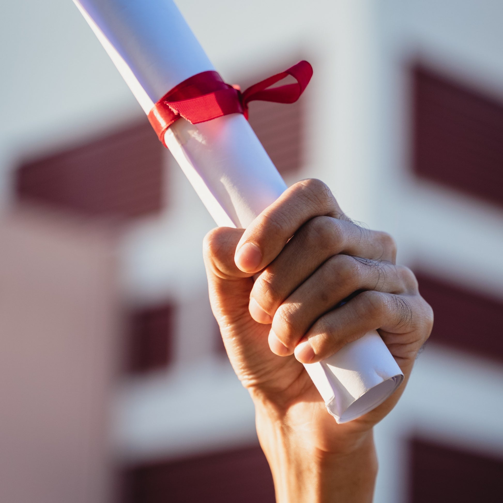 <Male hand in front of a blurred purple and white university college building holding in the air a rolled up degree certificate qualification tied with a red ribbon. Credit> AdobeStock_406407277