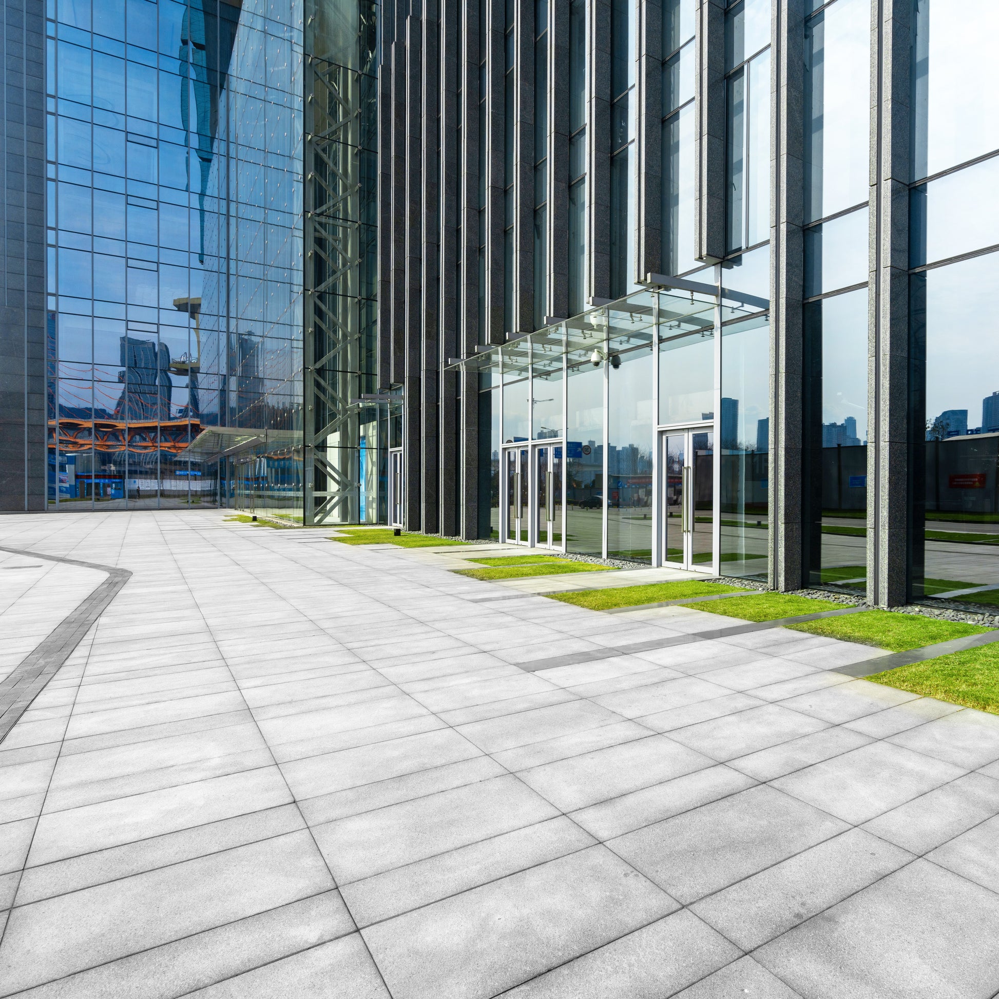 <Stone paved area with symmetric patterns in front of a modern glass L shaped office building with small green areas. Credit> AdobeStock_414022077