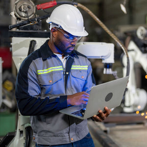 <Maintenance Enginneer wearing blue overall, white hardhat and safety glasses analyzing a white robotic arm packing unit with a laptop computer. Credit> AdobeStock_438287786