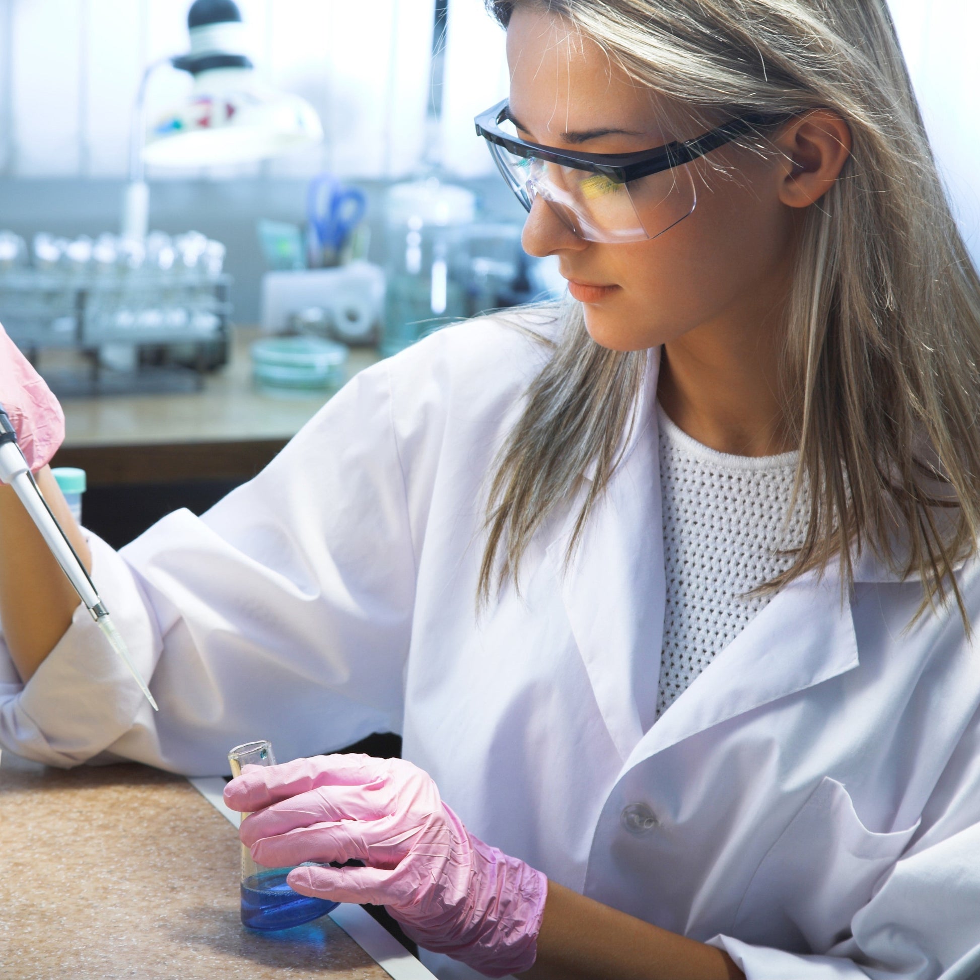 <Female laboratory analyst wearing a white lab coat, pink gloves and safety specs, pipetting a solution into a blue liquid in a glass conical flask. Credit> AdobeStock_44142293