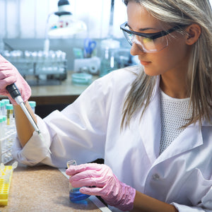 <Female laboratory analyst wearing pink gloves pipetting into a conical flask of clear blue liquid. Credit> AdobeStock_44142293