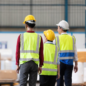 <Senior warehouse manager on a walkaround with two operators, all wearing fluorescent jackets and hard hats, his white, theirs yellow, with their backs to the camera. Credit> AdobeStock_449672578